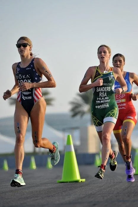 Three female athletes running on a road race course lined with green cones, with a palm tree and blue sky in the background.