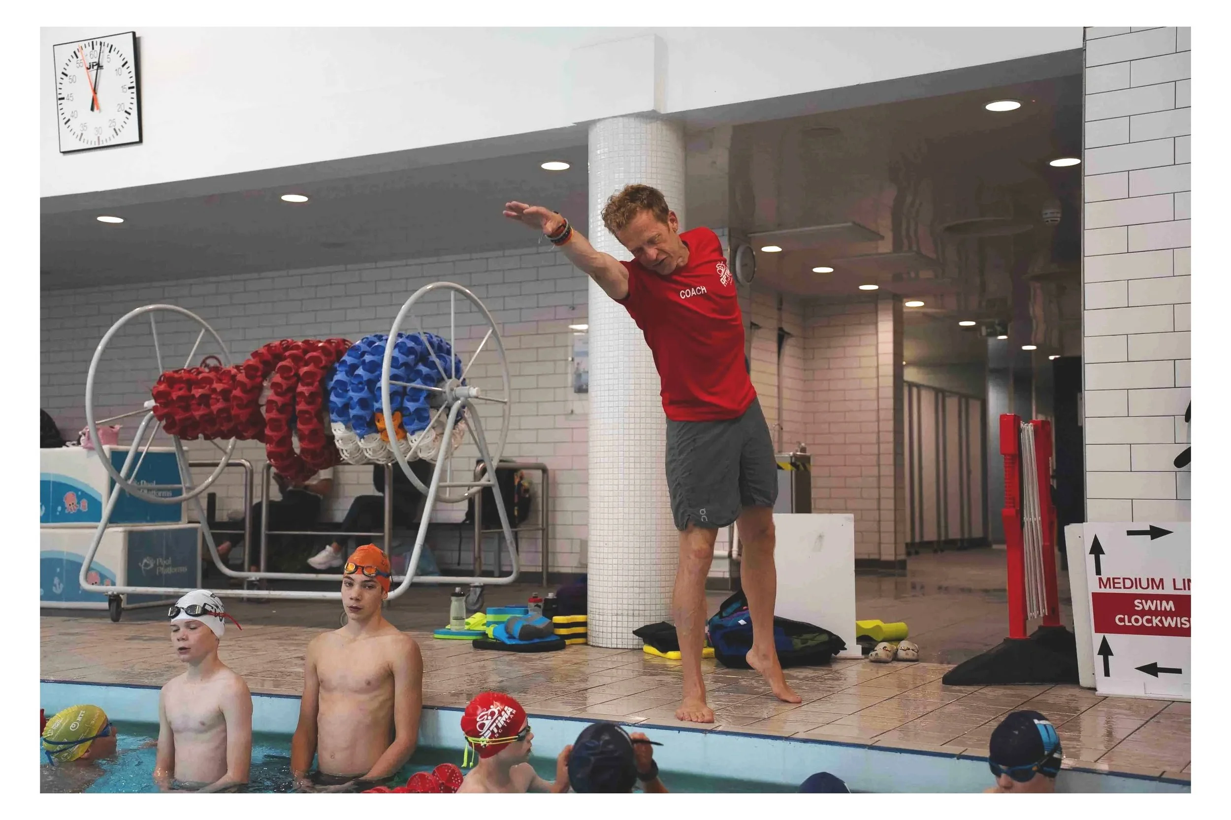 Swim coach demonstrating stretching exercise to young swimmers in indoor swimming pool area.