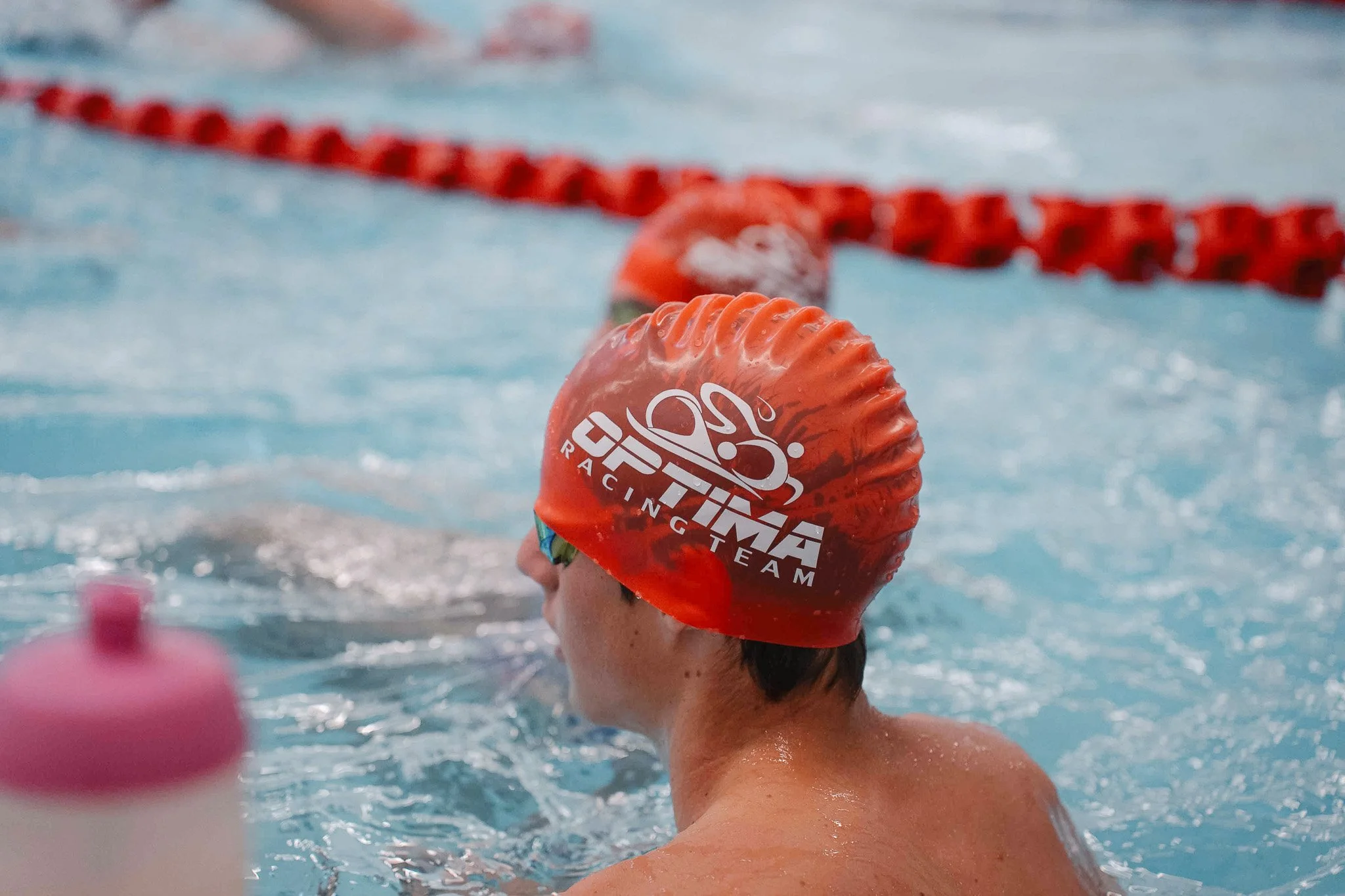 Swimmer wearing a red cap with the logo 'Optima Racing Team' in a swimming pool.