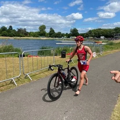 A woman in red athletic gear and a red helmet running beside a black bike on a paved path near a body of water during a triathlon event.