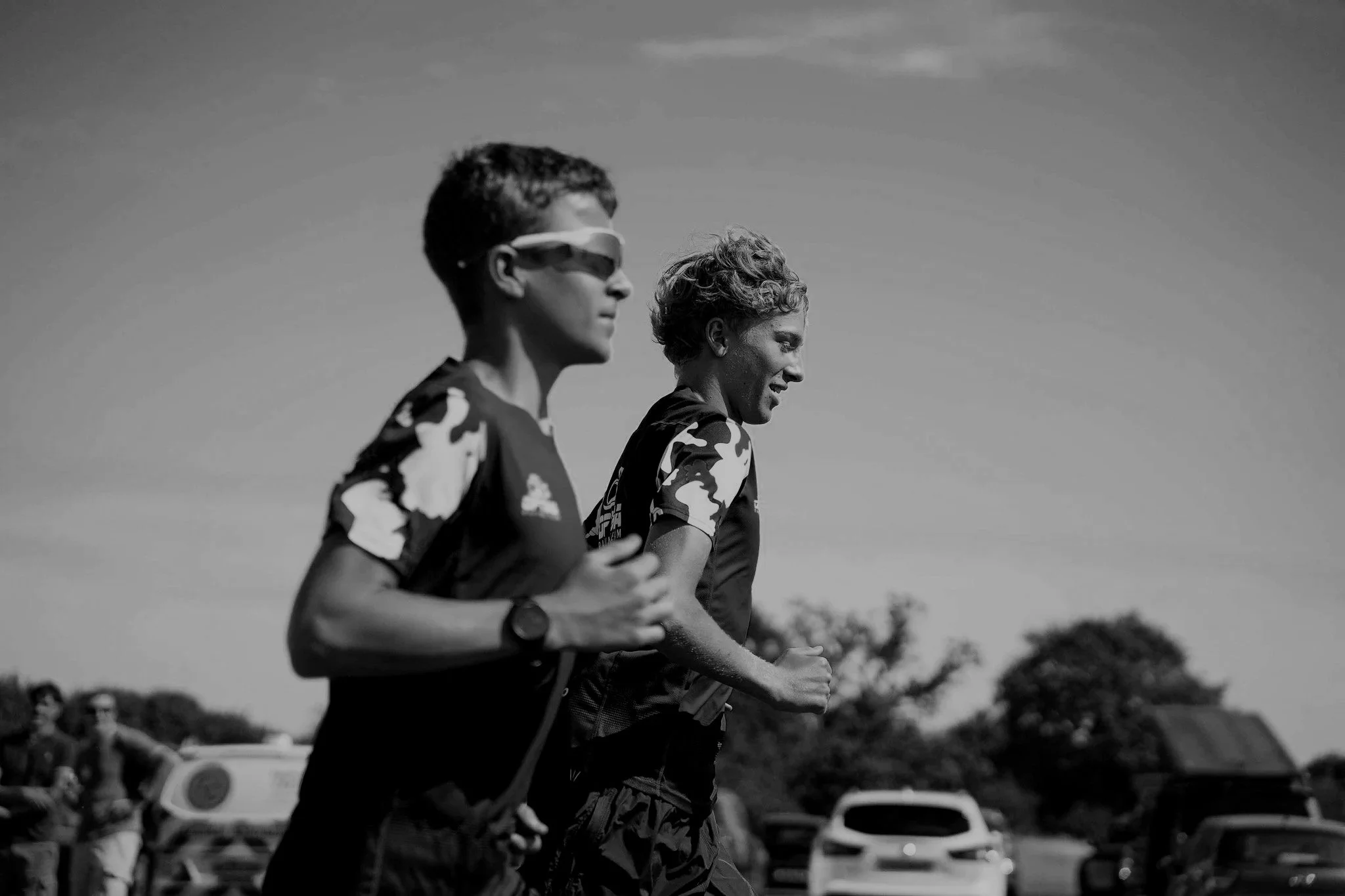 Two runner athletes, a young man wearing sunglasses and a woman with short curly hair, running outdoors during daytime, with cars and trees in the background, in black and white.
