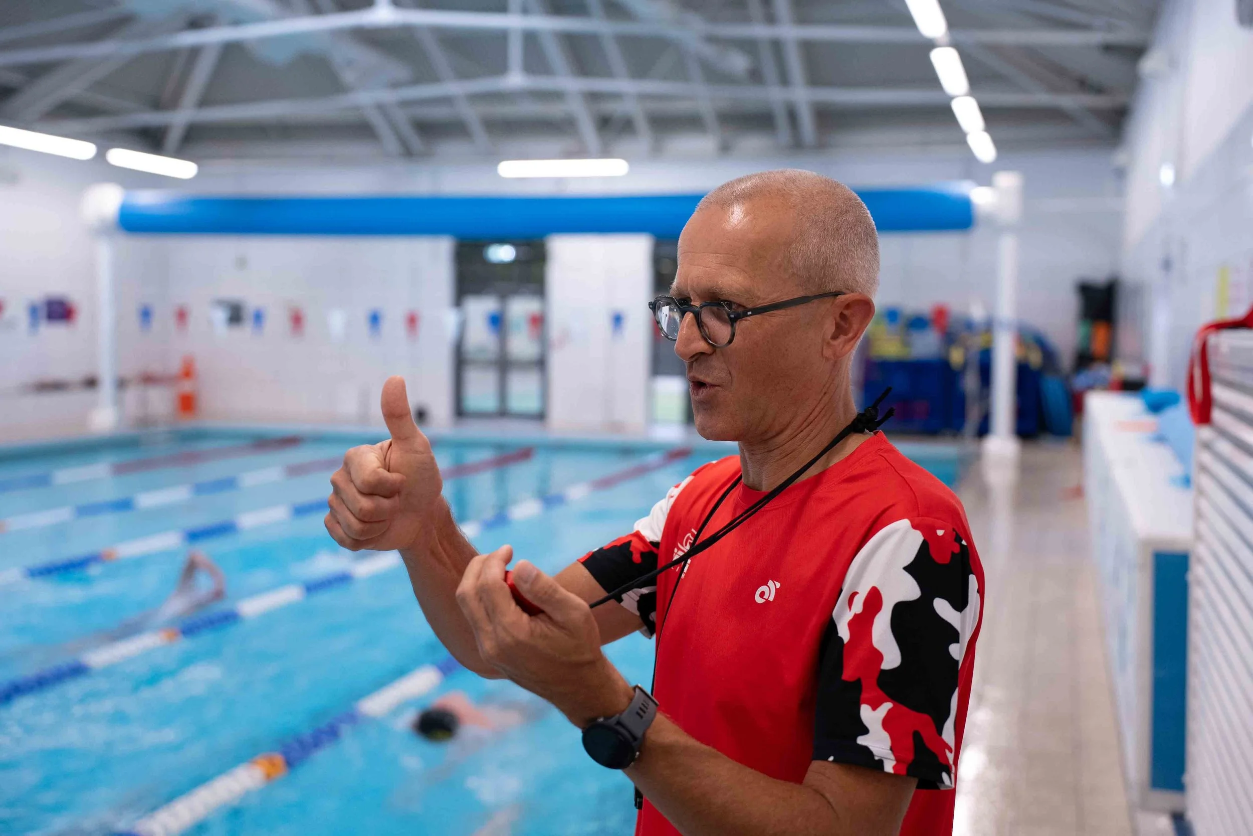Swim coach giving a thumbs-up gesture inside an indoor swimming pool area.