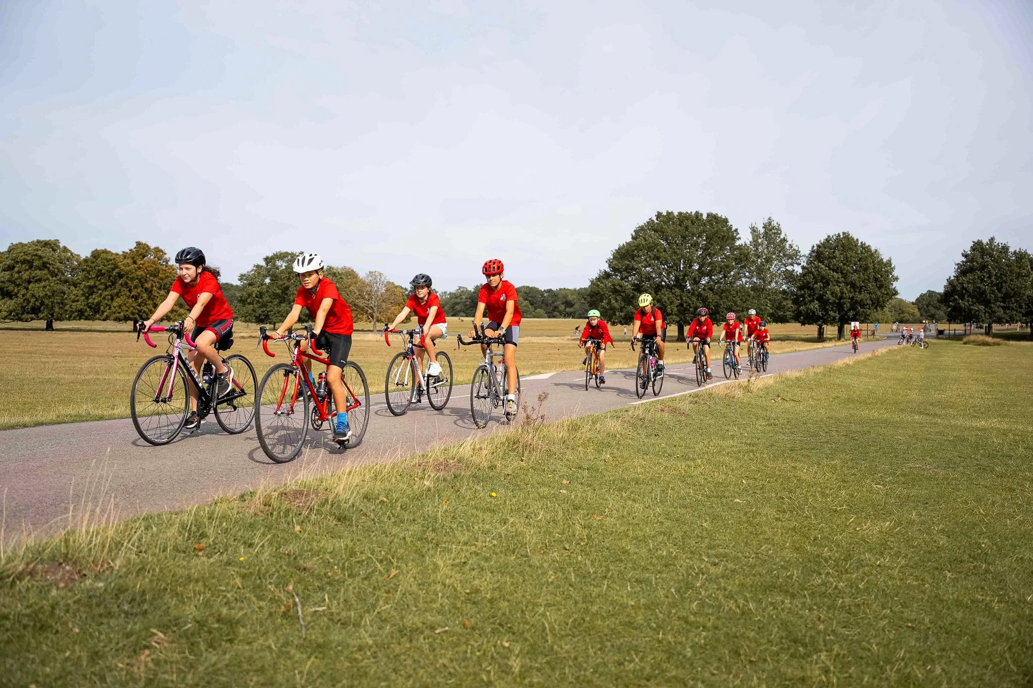 A group of children riding bicycles on a paved path through a park, all wearing helmets and red shirts.