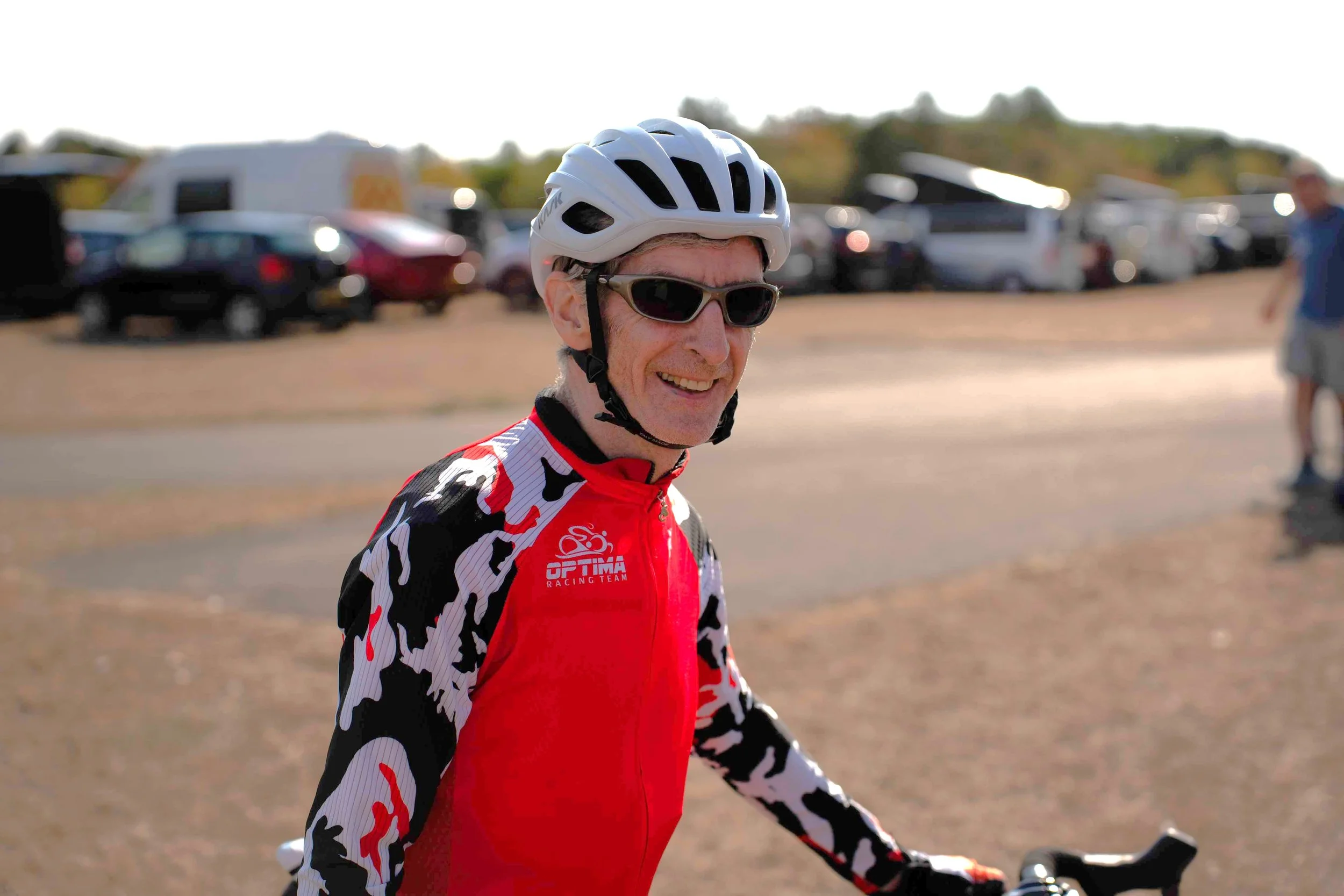 A man wearing a cycling helmet, sunglasses, and a red, black, and white cycling jersey smiling while standing next to his bicycle in a parking lot with cars and an RV in the background.
