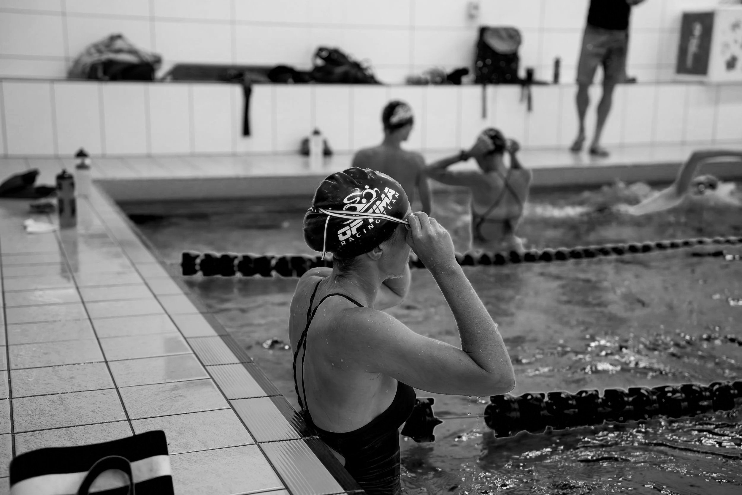 Swimmers preparing for practice or competition in an indoor swimming pool, with some adjusting swim caps and others in the water.