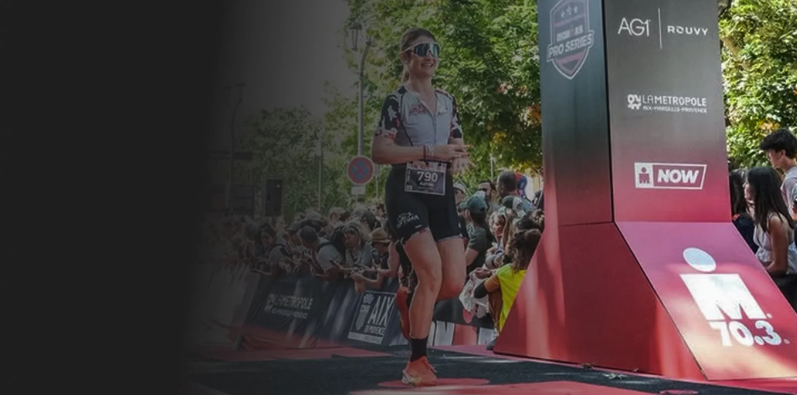 A woman finishing a race at the marathon, wearing black athletic gear, sunglasses, and a race bib, crossing the finish line with spectators in the background.