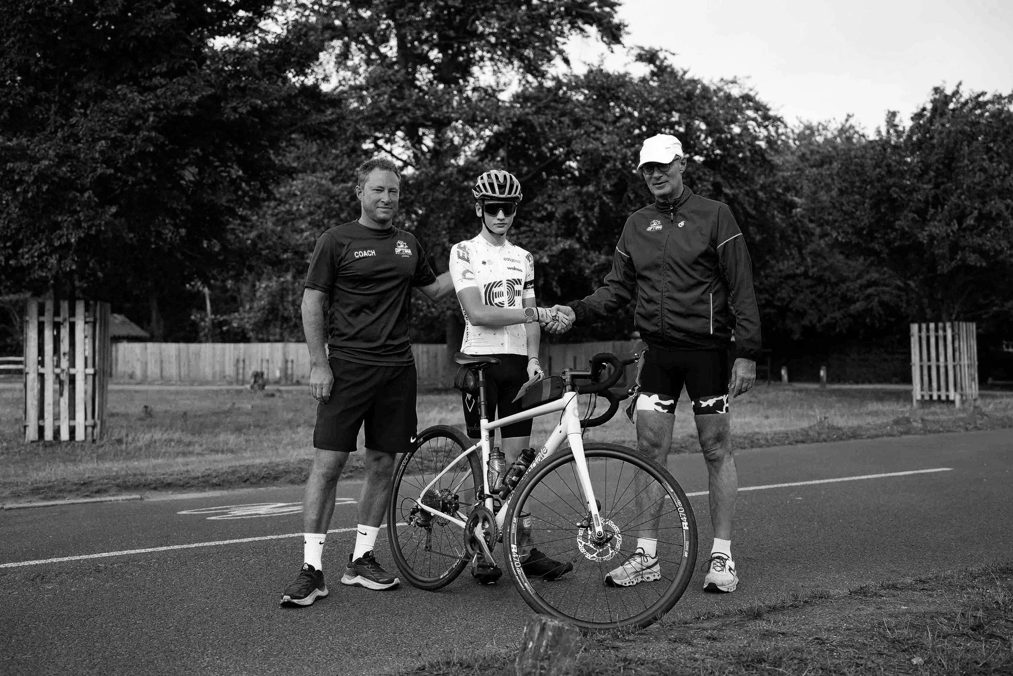 Three people standing outdoors, with the person in the middle holding a bicycle. The person on the left and right are shaking hands with the cyclist. The person on the left is wearing athletic clothing with a shirt that says 'coach,' and the person on the right is wearing a jacket, cycling shorts, and a cap. There are trees and a wooden fence in the background.
