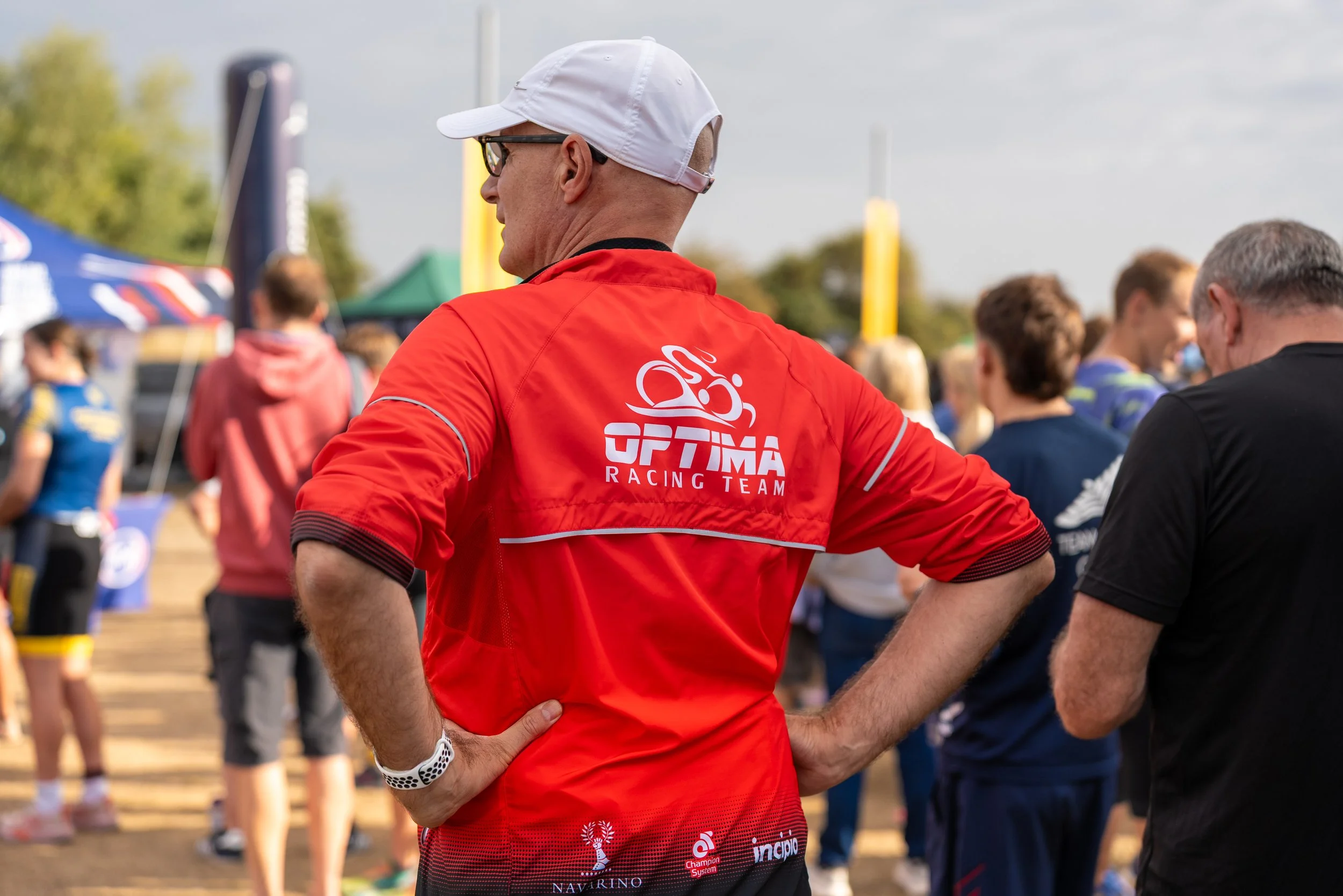 A man wearing a red Optima Racing Team shirt, white cap, and glasses, standing outdoors at a sporting event with other people and tents in the background.