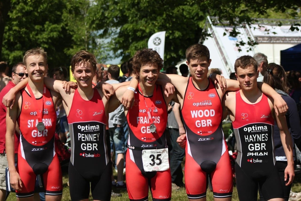Five young Optima Racing triathletes wearing red and black racing suits with various sponsor logos, standing outdoors with their arms around each other's shoulders, smiling after a race, with medals around their necks and a crowd in the background.