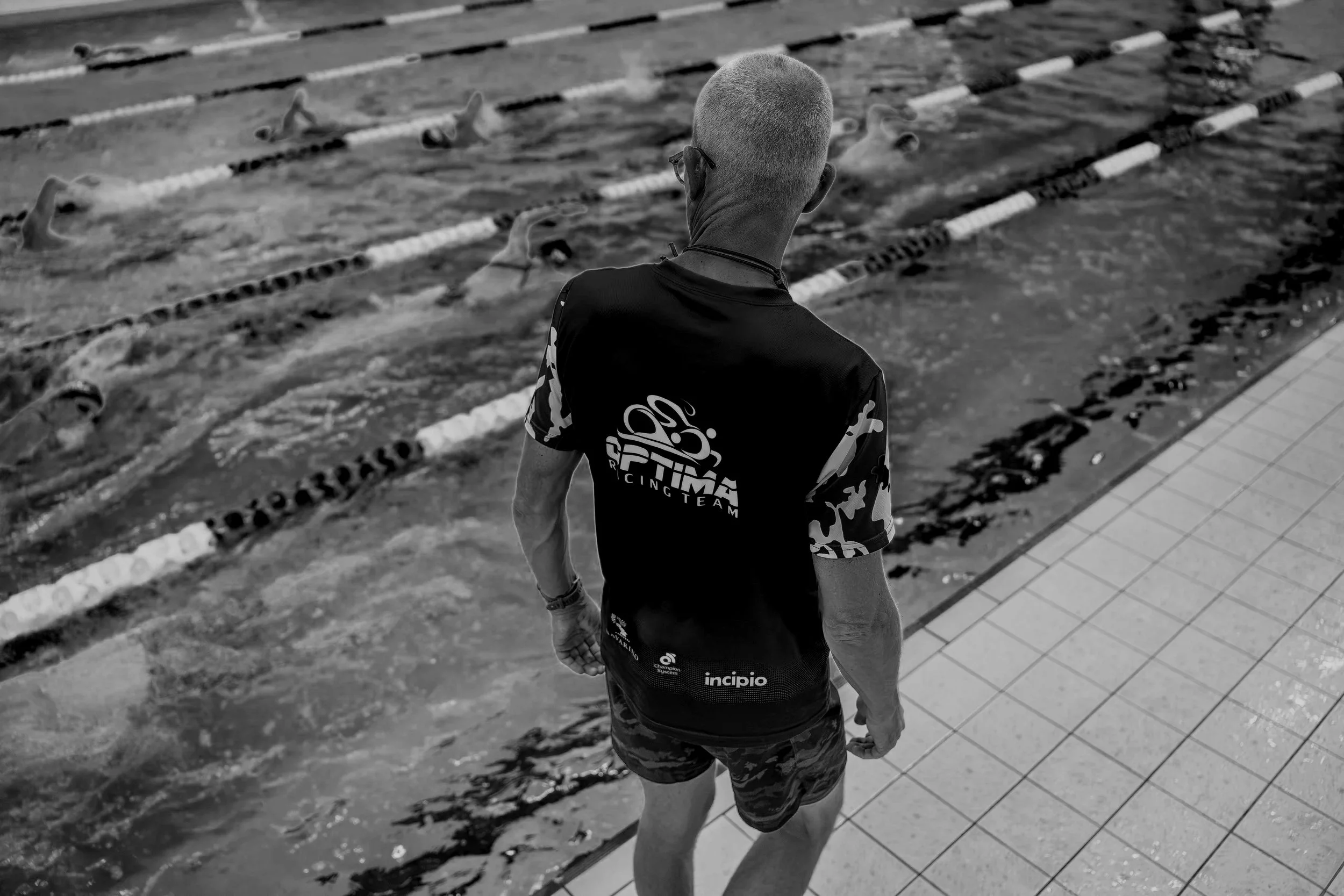 A person standing at the edge of a swimming pool, wearing a sports shirt with 'FITA Racing Team' text on the back and camouflage shorts, looking at the pool.