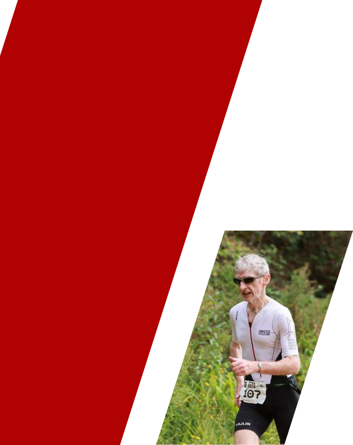 An elderly woman running outdoors, wearing sunglasses, a white athletic shirt, and black shorts, with green foliage in the background.