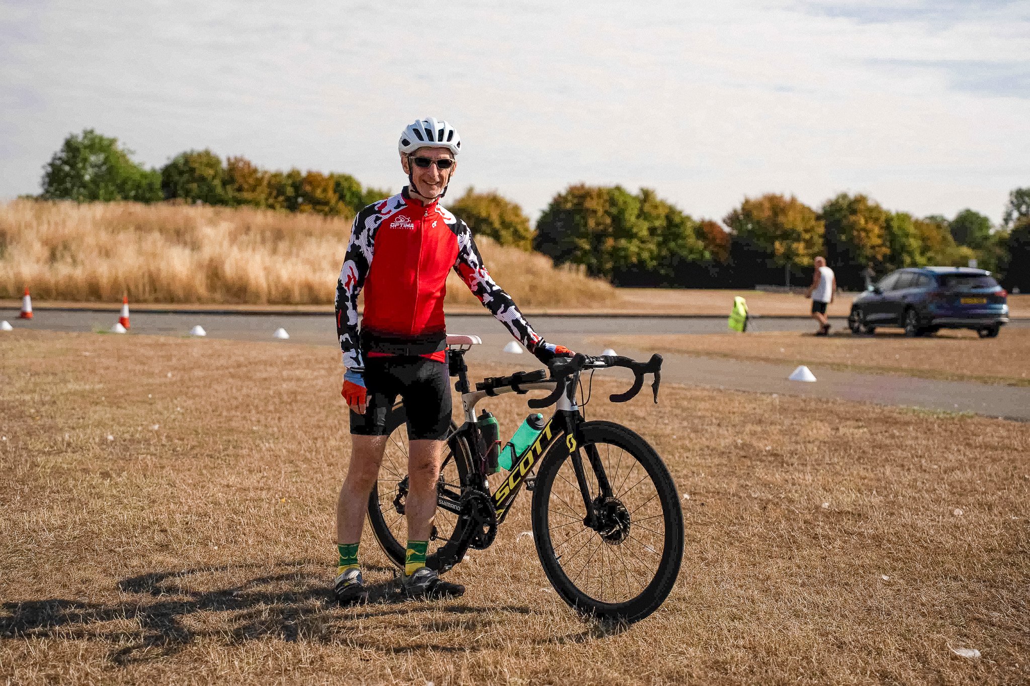 A man in cycling gear standing next to a black and yellow Scott road bicycle on a grassy field, with a few people and a parked car in the background.