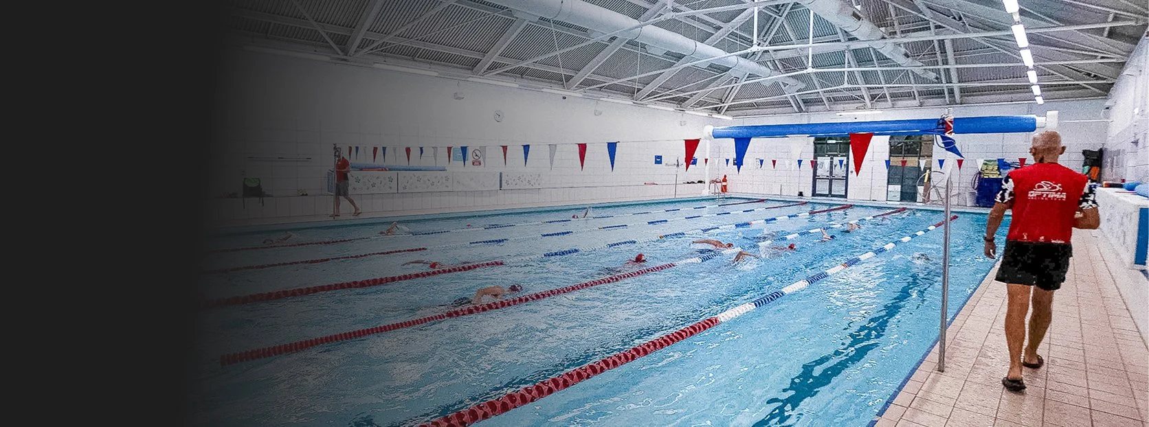 Indoor swimming pool with swimmers practicing laps, red and blue pennant flags hanging above the pool, and a coach walking along the poolside.