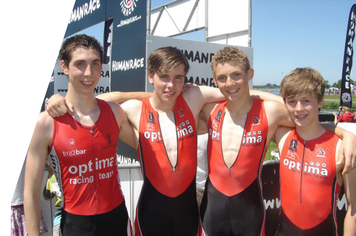 Four young male Optima Racing athletes in red racing suits with their arms around each other, smiling at the camera, outdoors at a sporting event with banners and a blue sky in the background.