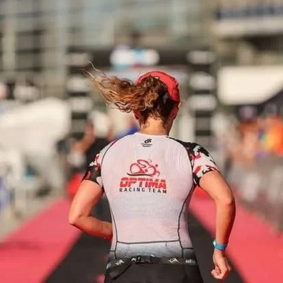 Female athlete running outdoors with hair flowing, wearing a white and pink Optima Racing Team shirt and a red cap.