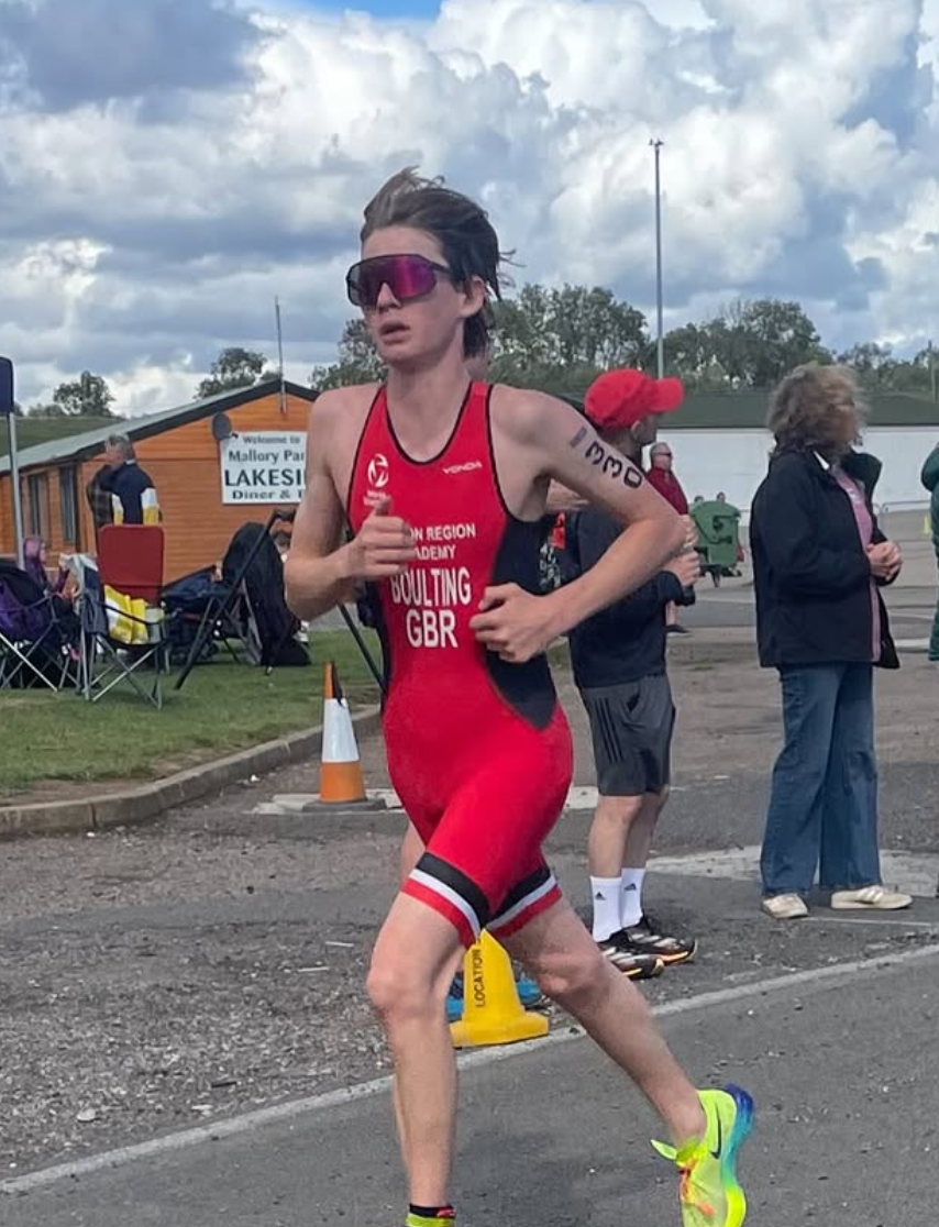 A female athlete running in a race, wearing a red and black triathlon suit, sunglasses, and neon yellow running shoes, with people and a building in the background.