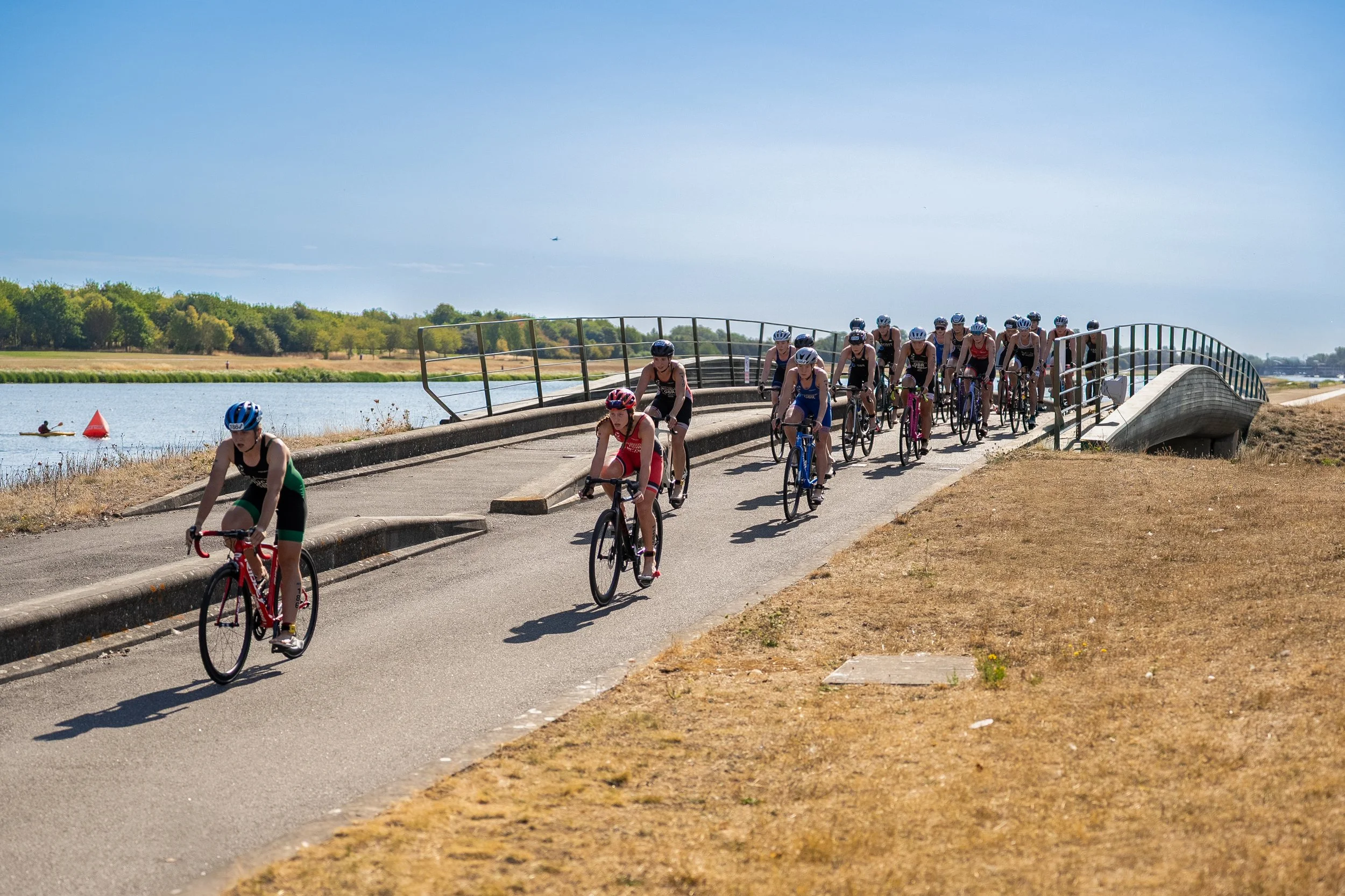 Group of Optima Racing athletes participating in a biking event on a bridge near a body of water, with trees in the background and a partly cloudy sky.