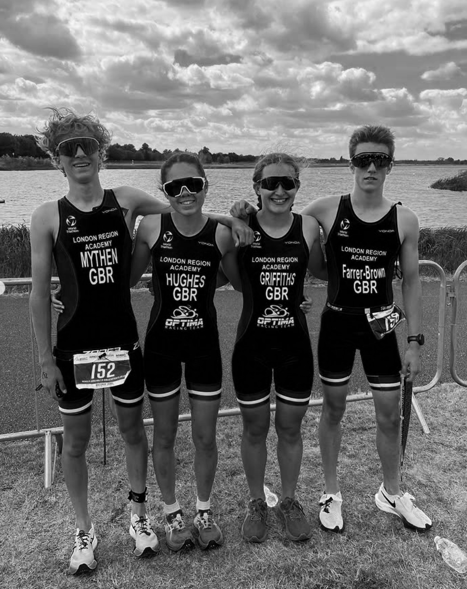 Four young athletes in black uniforms with goggles, standing outdoors near a body of water, smiling and posing for a team photo, with a cloudy sky overhead.
