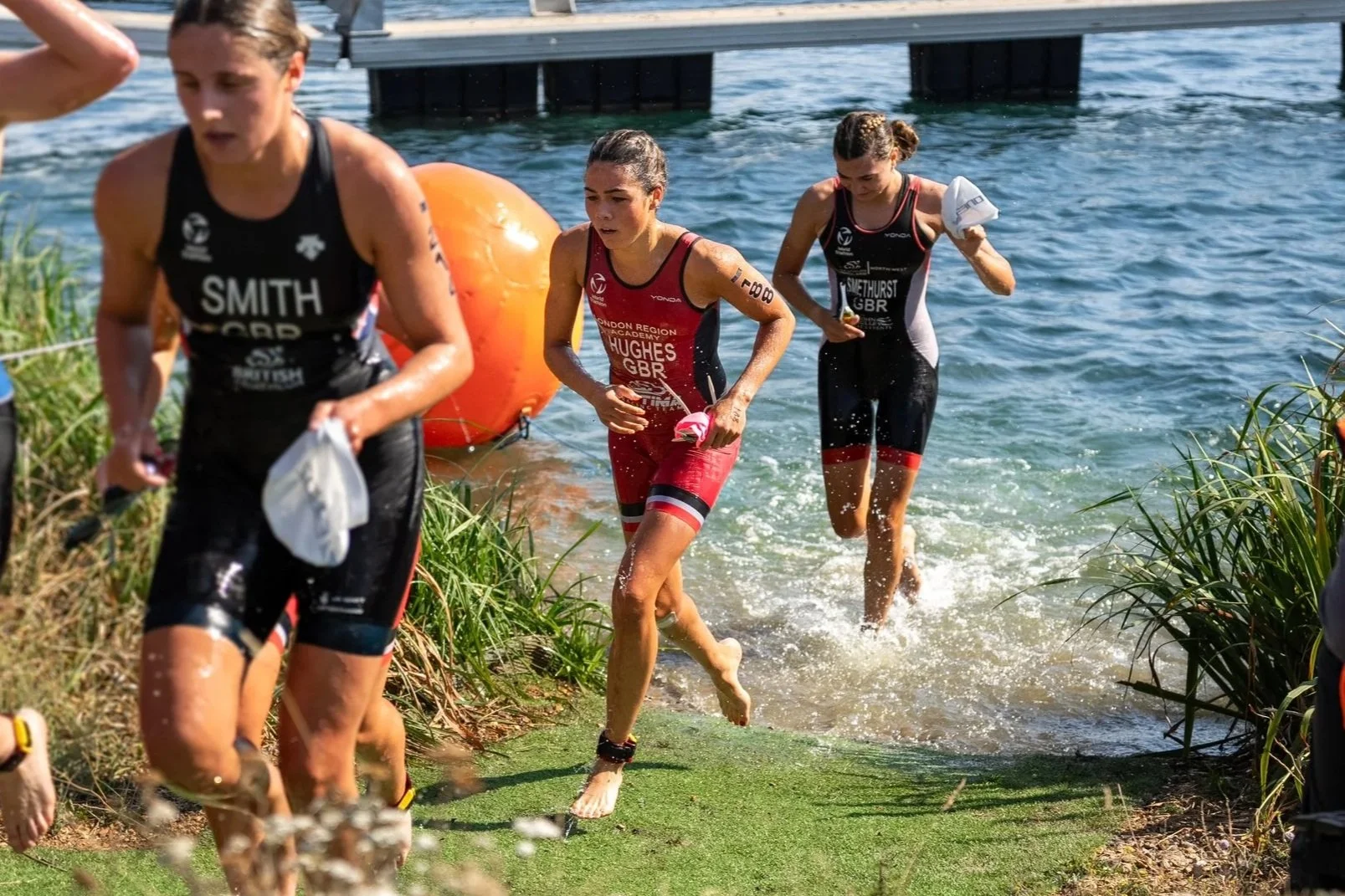 Triathlon participants exiting the water onto the shore, wearing wetsuits and swim caps, with a large orange buoy in the water and a dock in the background.