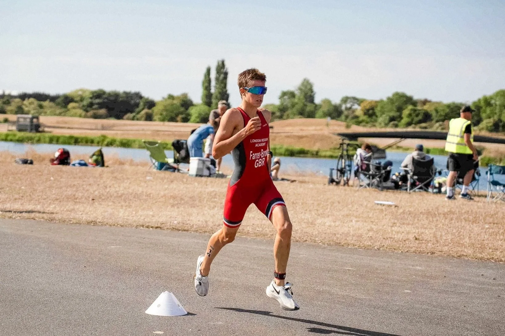 A male triathlete in a red and black suit running on a paved road near a body of water, with spectators and volunteers in the background, some sitting in chairs and others standing, during a race.