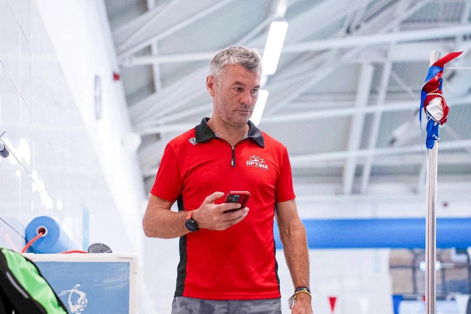 Coach James Beckinsale in a red and black sports shirt standing in an indoor sports facility, looking at his smartphone.