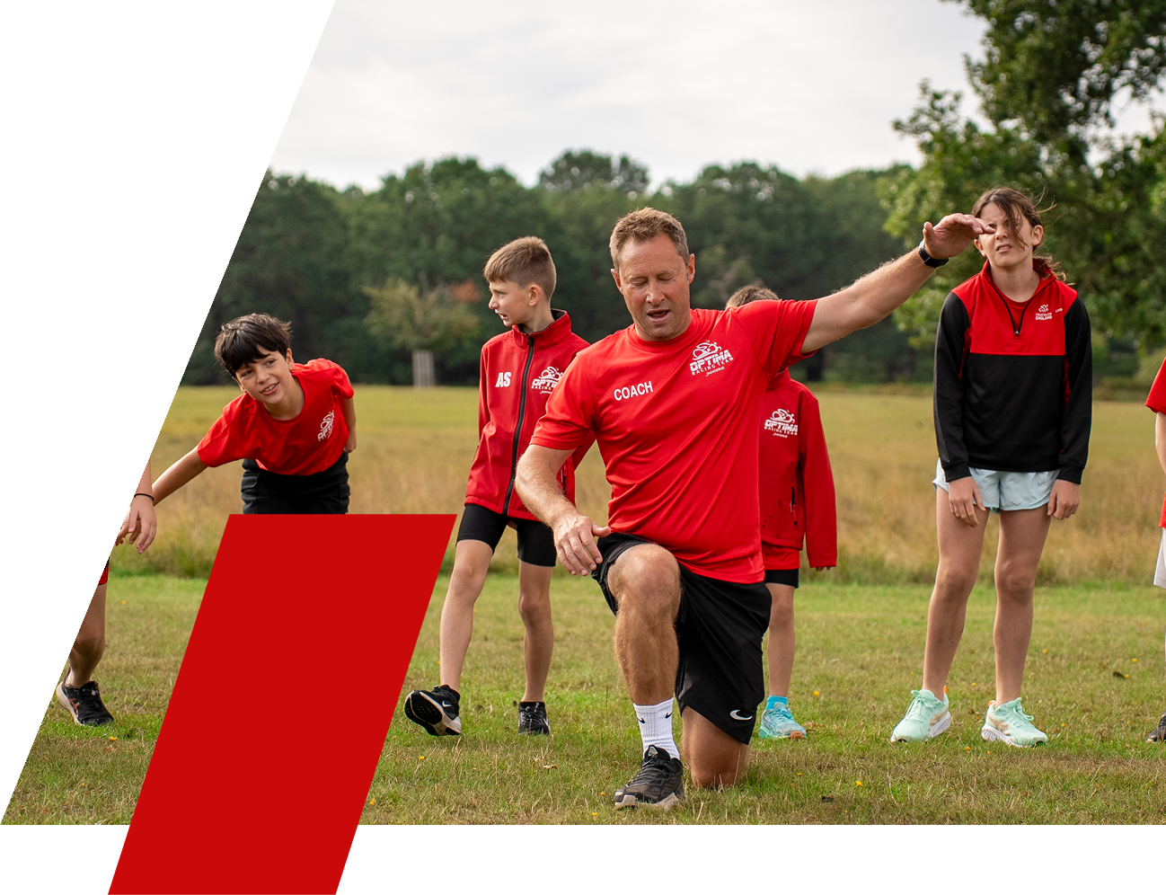 Coach Mark Klein kneeling on one knee giving instructions to children during an outdoor training or sports practice in a grassy field with trees in the background, all wearing red sports shirts.
