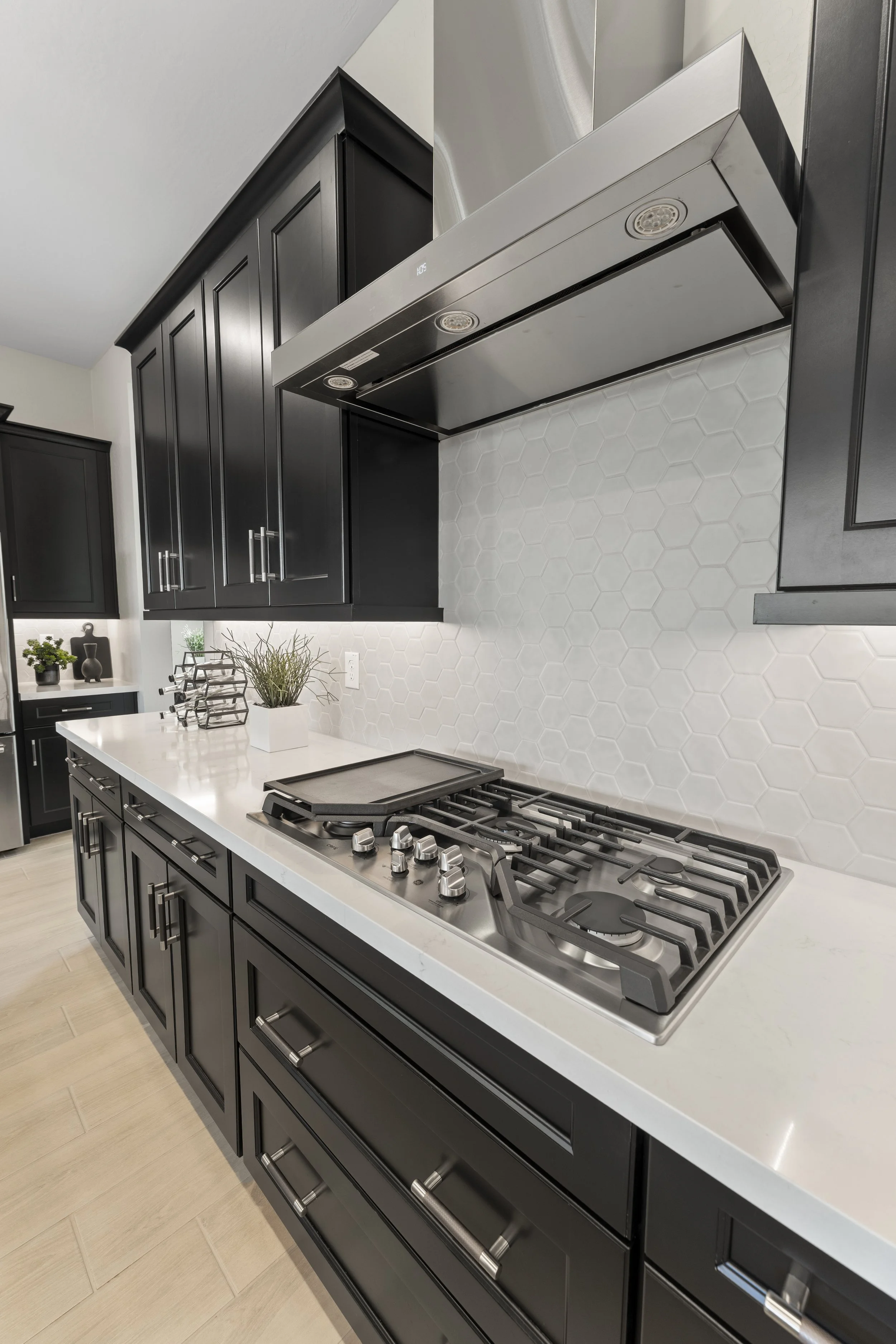 Modern kitchen with black cabinetry, white countertop, gas stove, stainless steel range hood, white hexagon tile backsplash, and small potted plant.