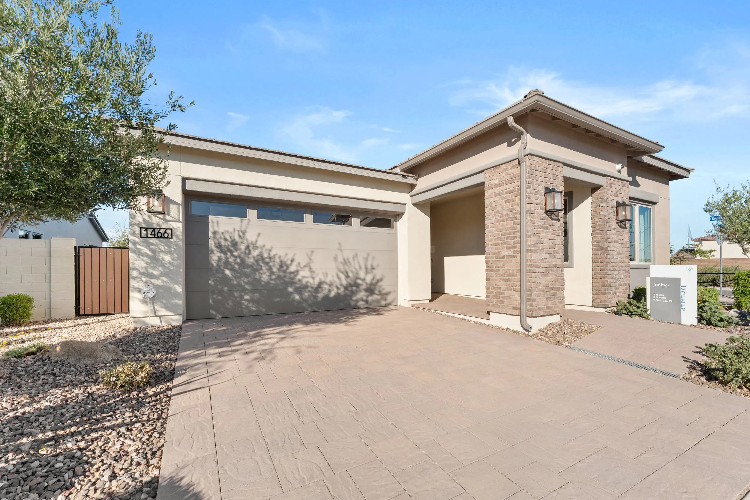 A modern, single-story house with a two-car garage, beige walls, brick accents near the entrance, and desert landscaping.