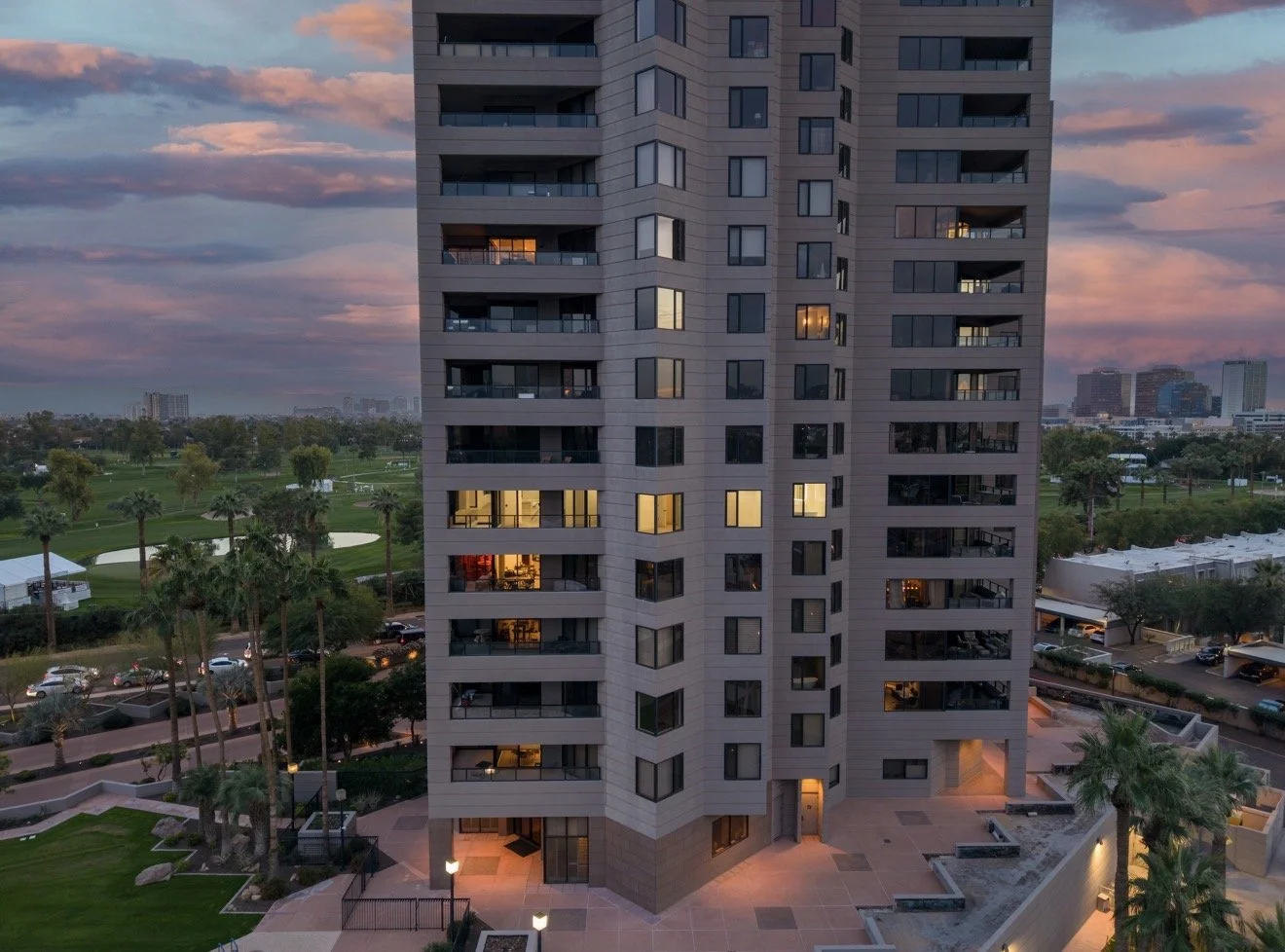 A tall residential apartment building at dusk with some lights on inside, urban background with other buildings and a green park with palm trees.