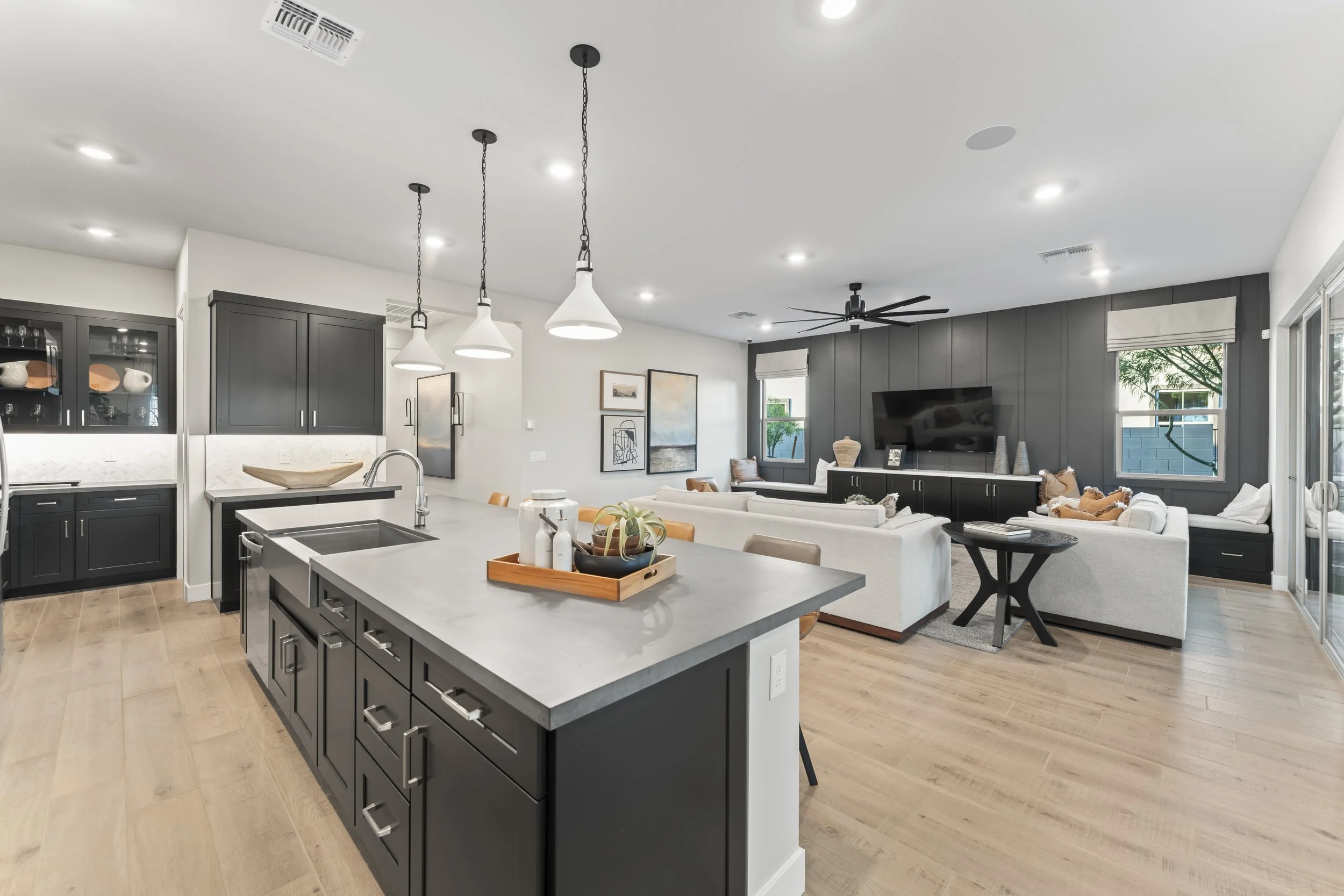 Open-concept living room and kitchen with black cabinetry, white countertops, light wood flooring, and contemporary furniture, with large windows and sliding glass doors.