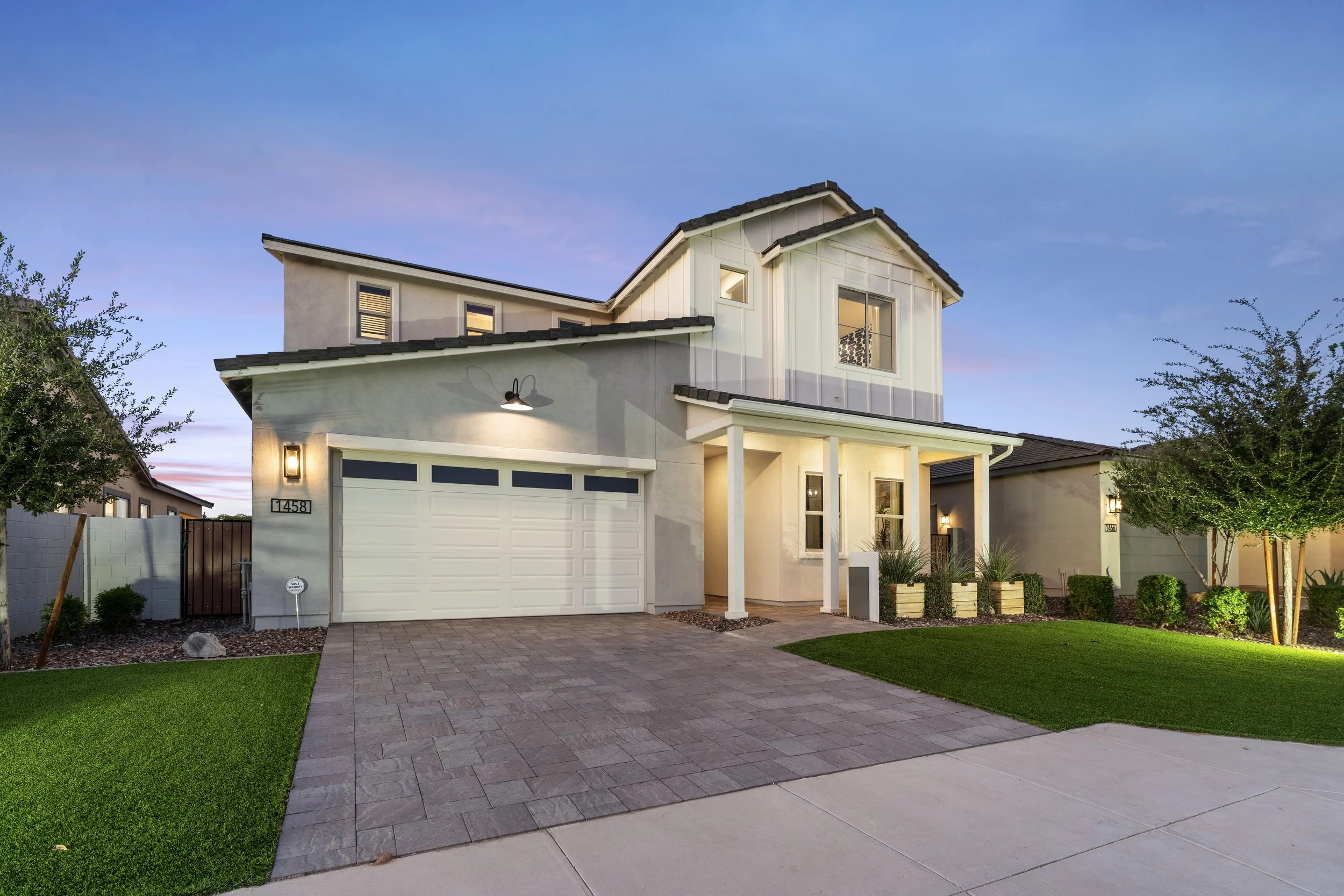 Modern two-story house with a landscaped front yard, driveway, garage, and porch, illuminated during twilight.