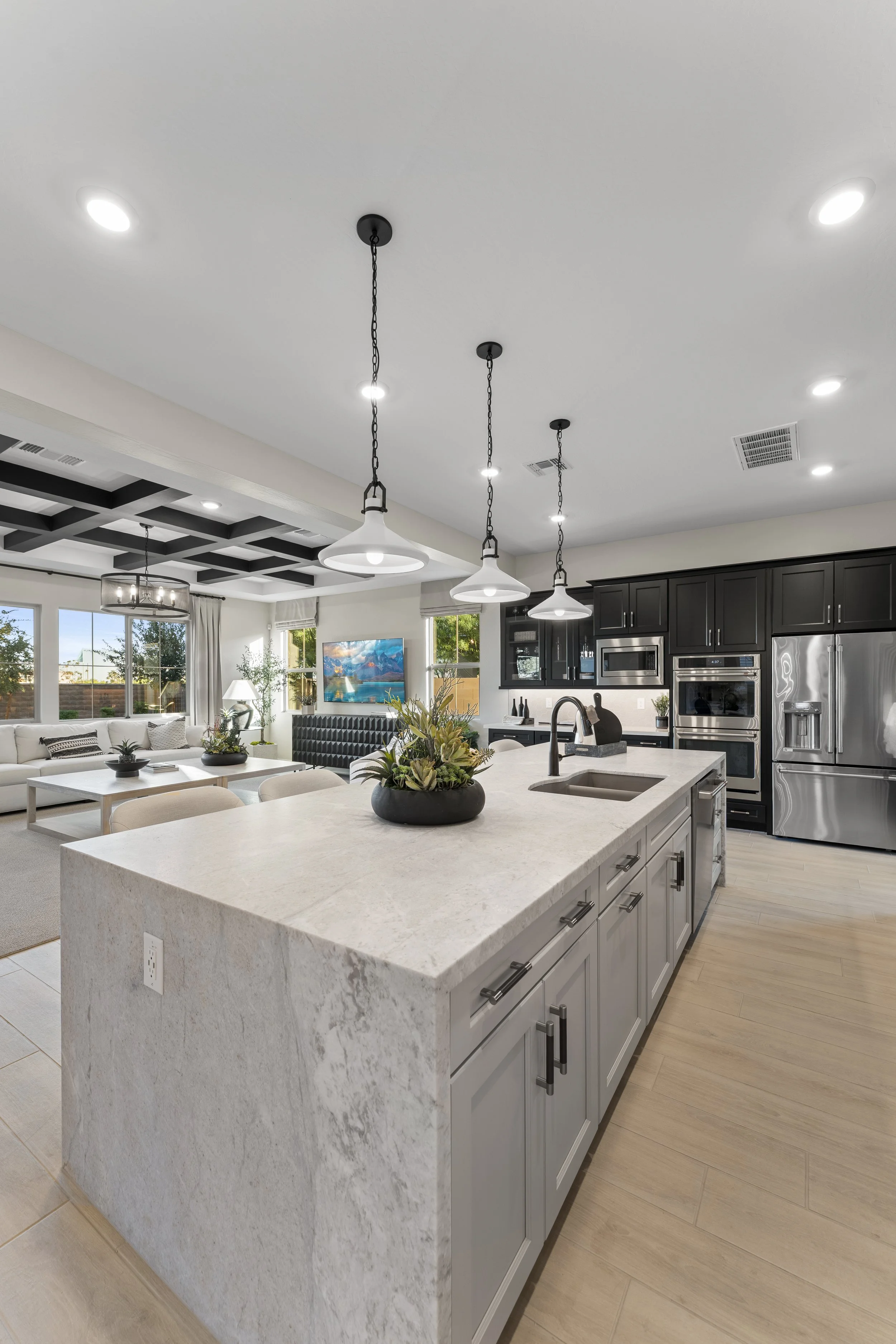 Open-concept kitchen with a large white island, black cabinets, stainless steel appliances, and a living room with a couch and television in the background.
