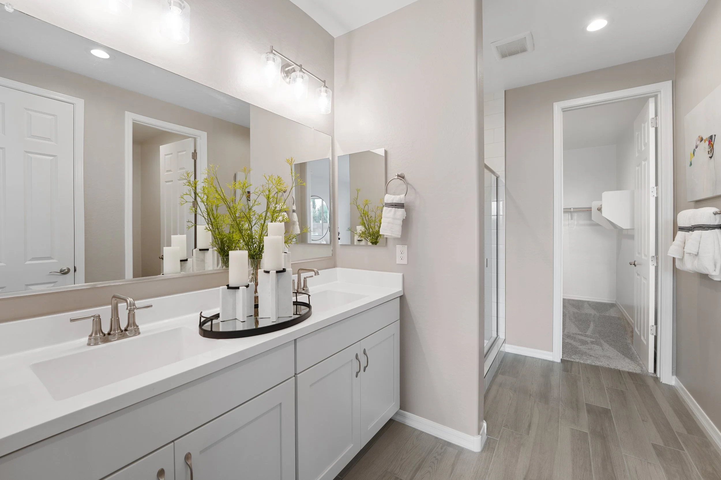 Modern bathroom with white vanity, large mirror, candles, green floral arrangement, and towels, with separate shower area and laundry space in the background.