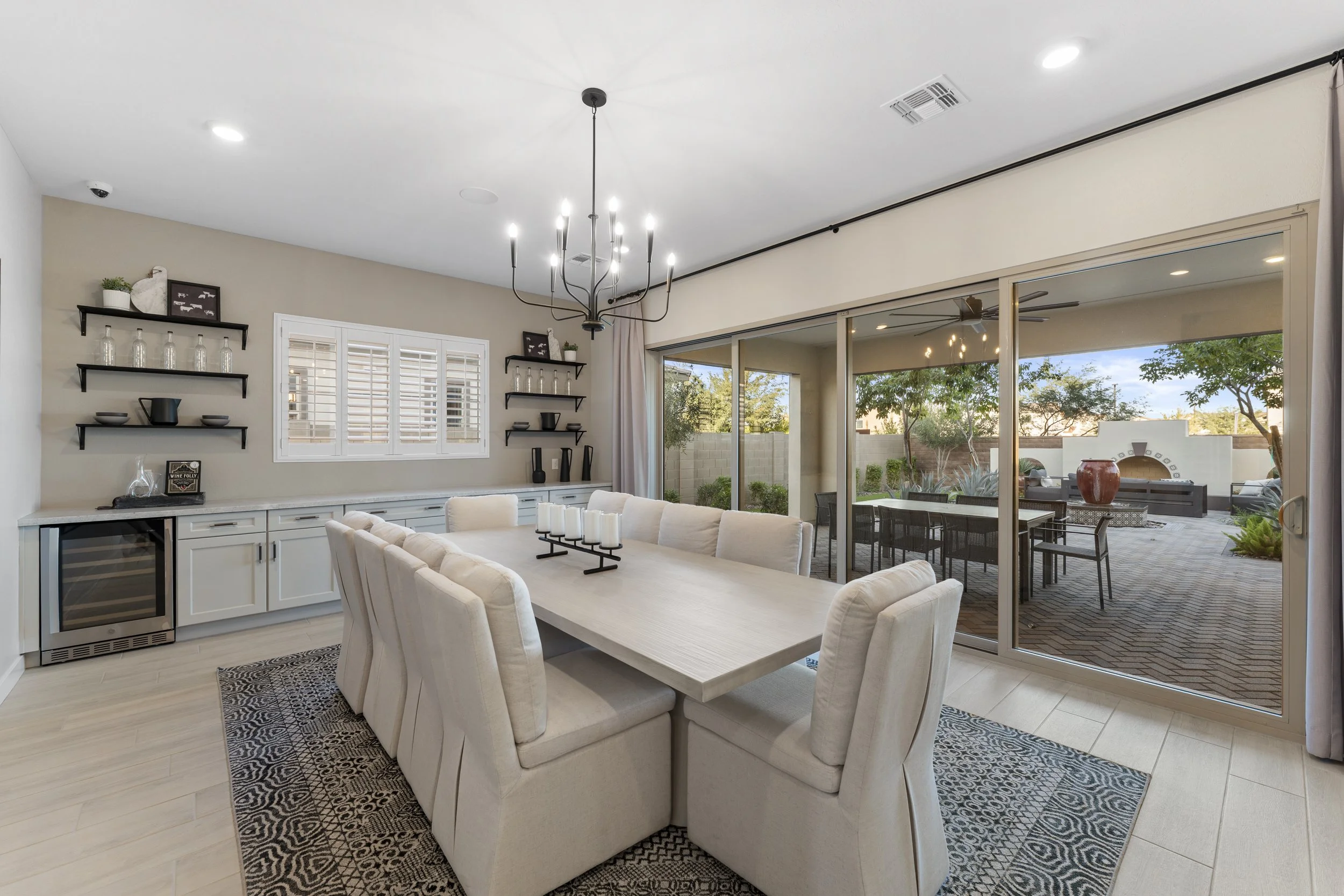 Dining room with a large white table and chairs, black floating shelves on beige walls, and a view of an outdoor patio with a fireplace and seating area through glass sliding doors.