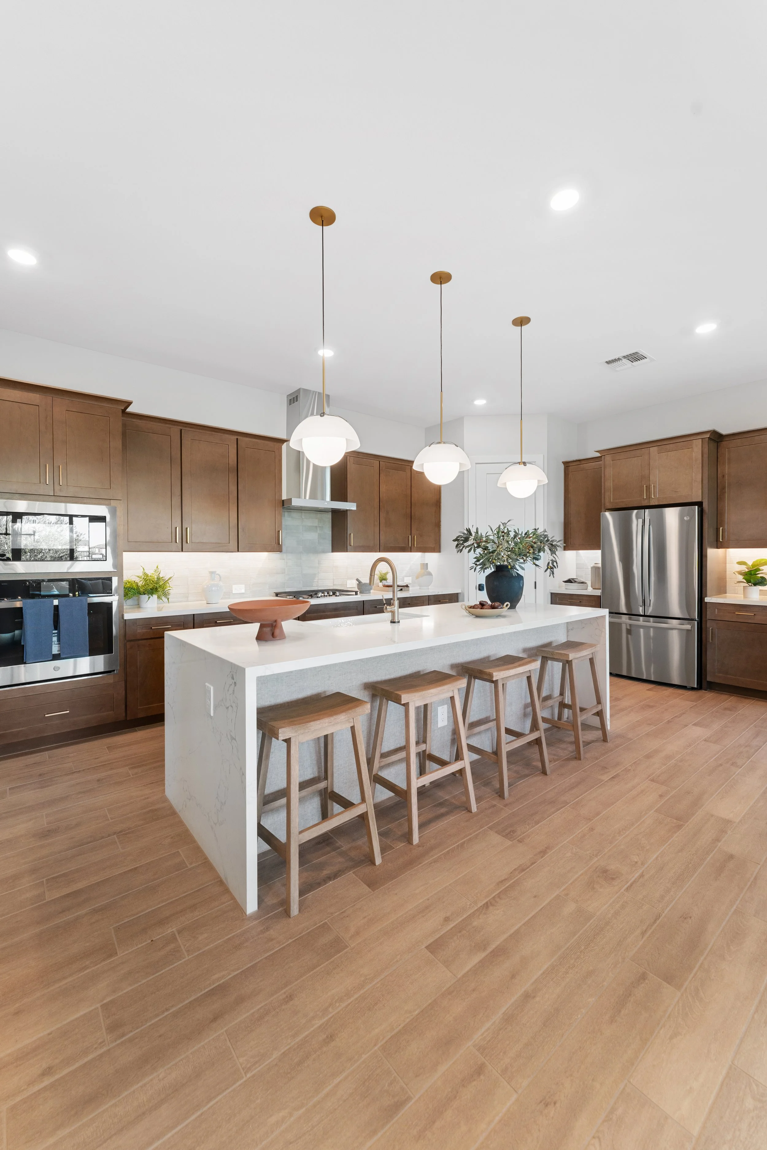 Modern kitchen with wooden cabinets, white island with seating, pendant lights, stainless steel refrigerator, and wooden flooring.