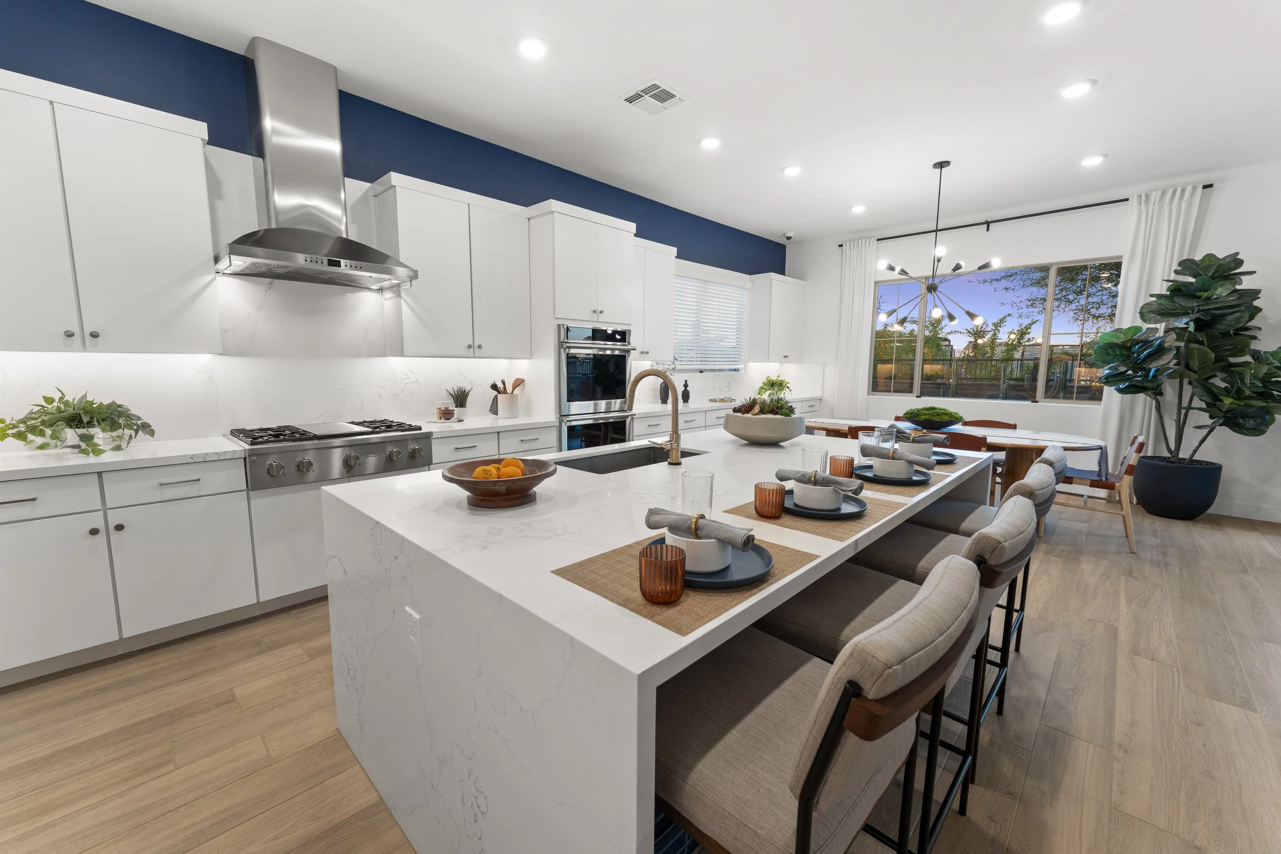 Modern kitchen with white cabinets, marble countertop, stainless steel appliances, and a large island with place settings. There is a dining area with a table and chairs near a large window, and a potted plant in the corner.