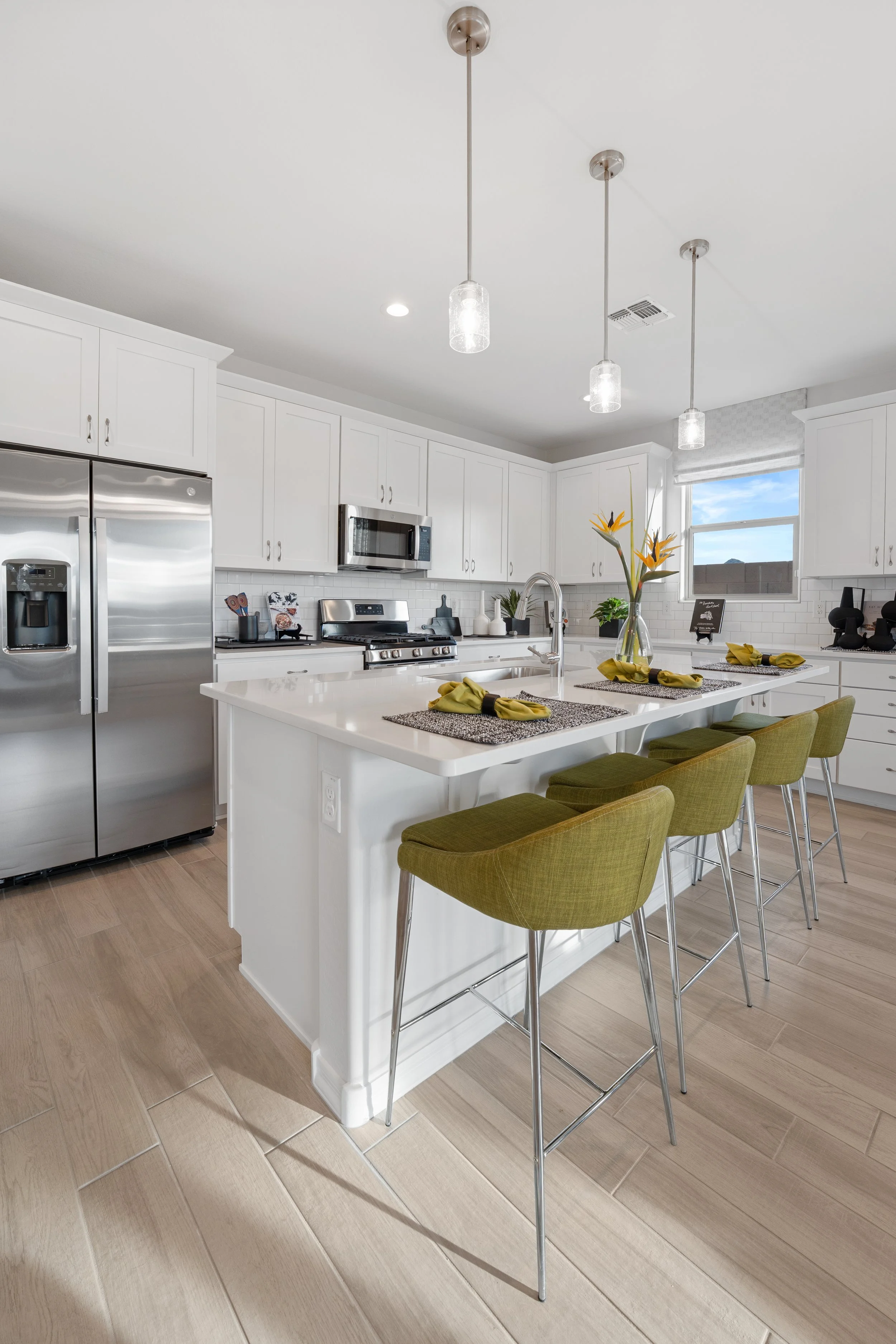 Modern kitchen with white cabinets, stainless steel appliances, and a white kitchen island with four green stools. The island has a vase with yellow flowers, and there are three pendant lights hanging from the ceiling. A window with a view of blue sky and clouds is in the background.