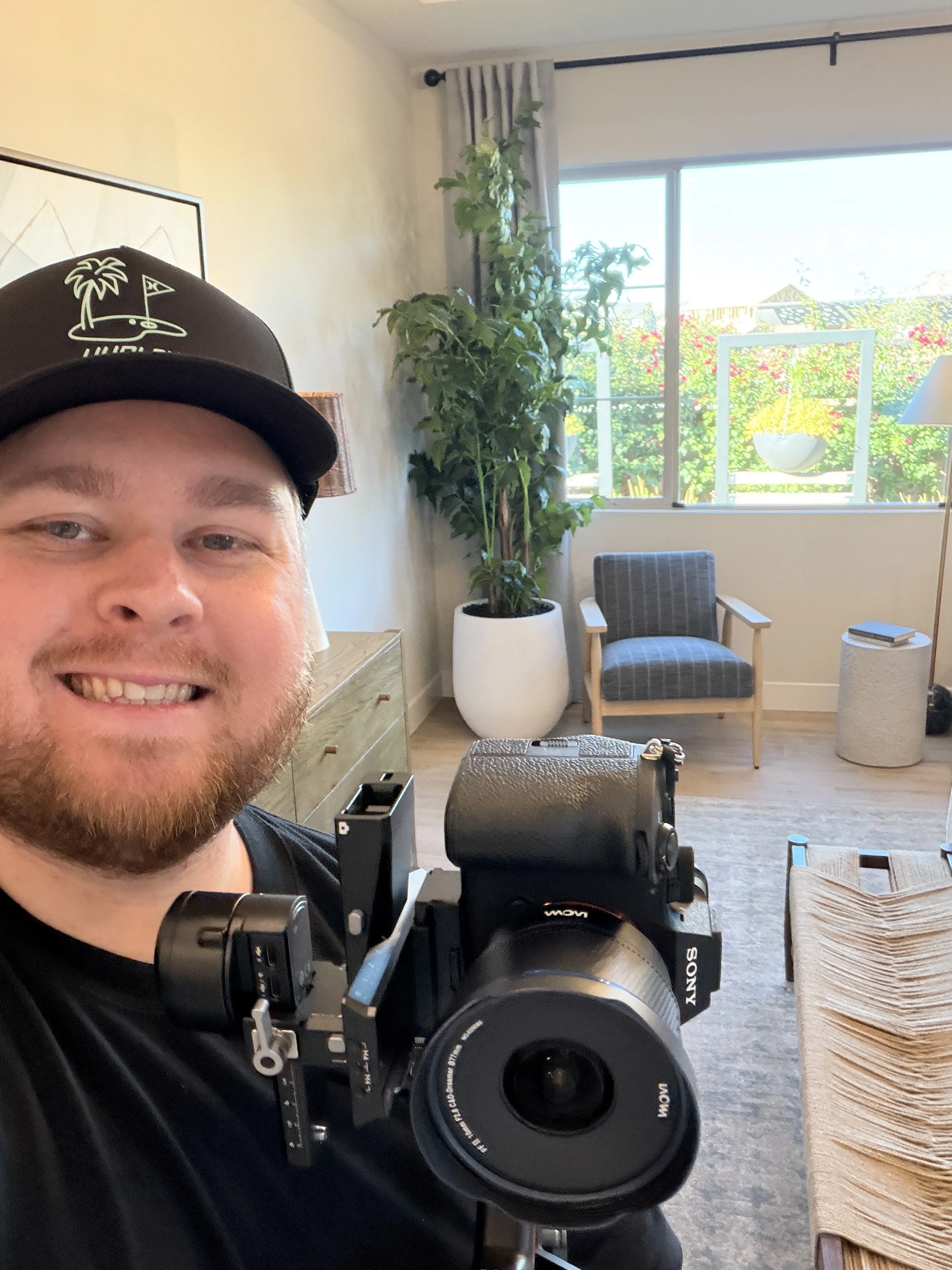 A smiling man taking a selfie in a bright living room with a professional camera. The room has a large window with a view of greenery, a potted plant, a chair, and a lamp.