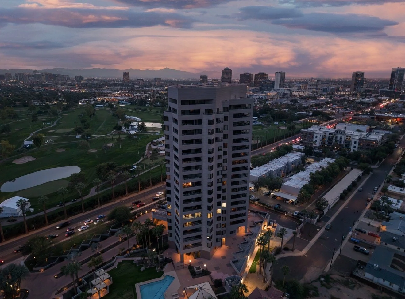 A tall modern apartment building with illuminated windows, surrounded by a well-lit courtyard, palm trees, and a swimming pool at sunset in an urban area with a golf course and city skyline in the background.