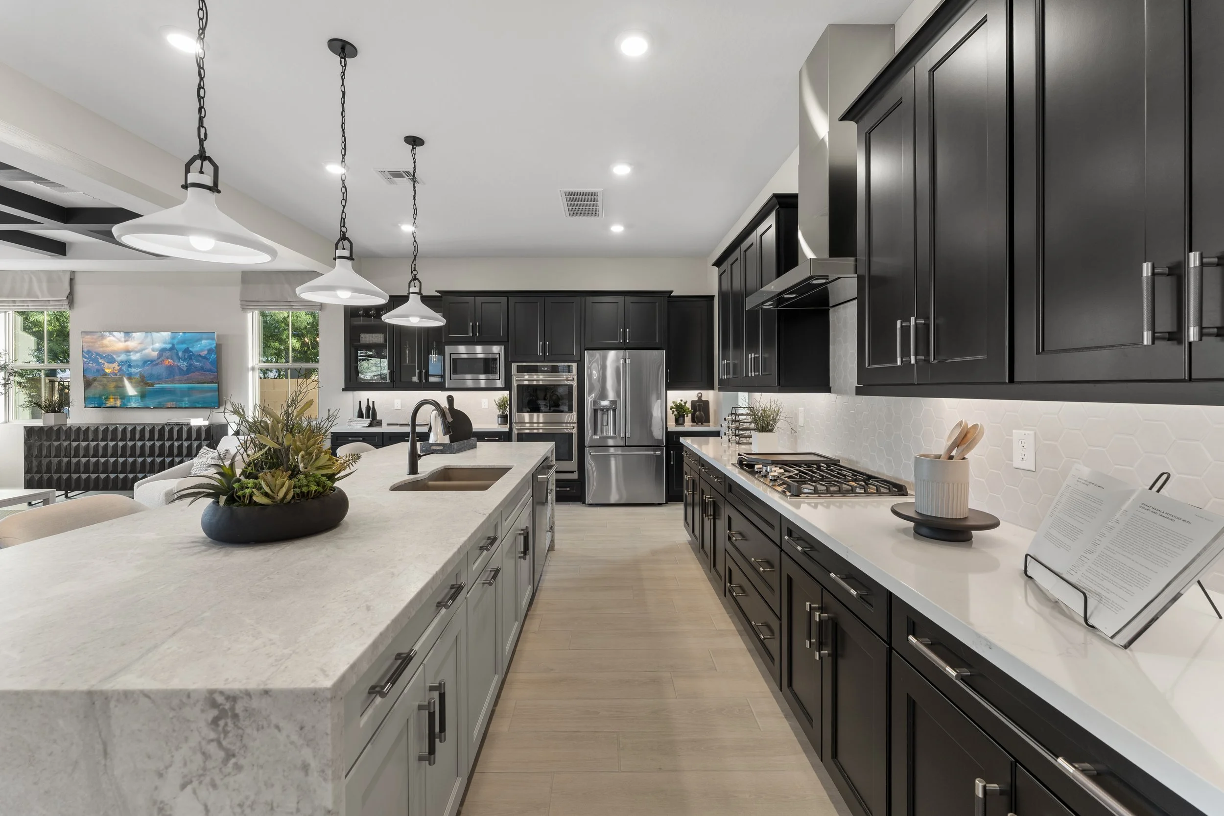 Modern kitchen with black cabinets, white countertops, and stainless steel appliances. Pendant lights hang above a large island with a sink. There is a gas stove, a coffee machine, and a book stand on the counters, with a living area and TV visible i