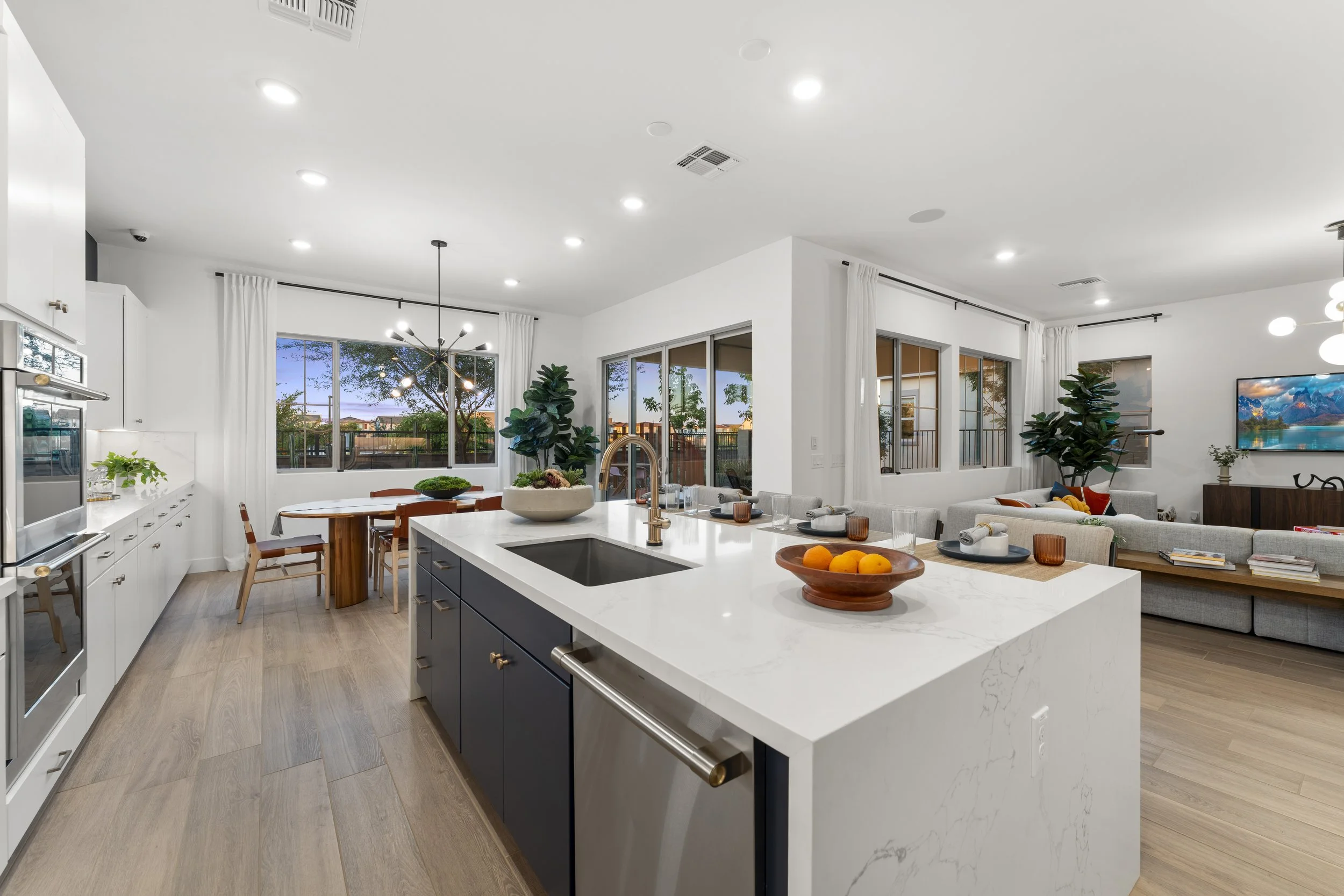Bright open-concept kitchen and living room with white cabinets, a kitchen island with a marble countertop, and large windows showing outdoor trees and evening sky.