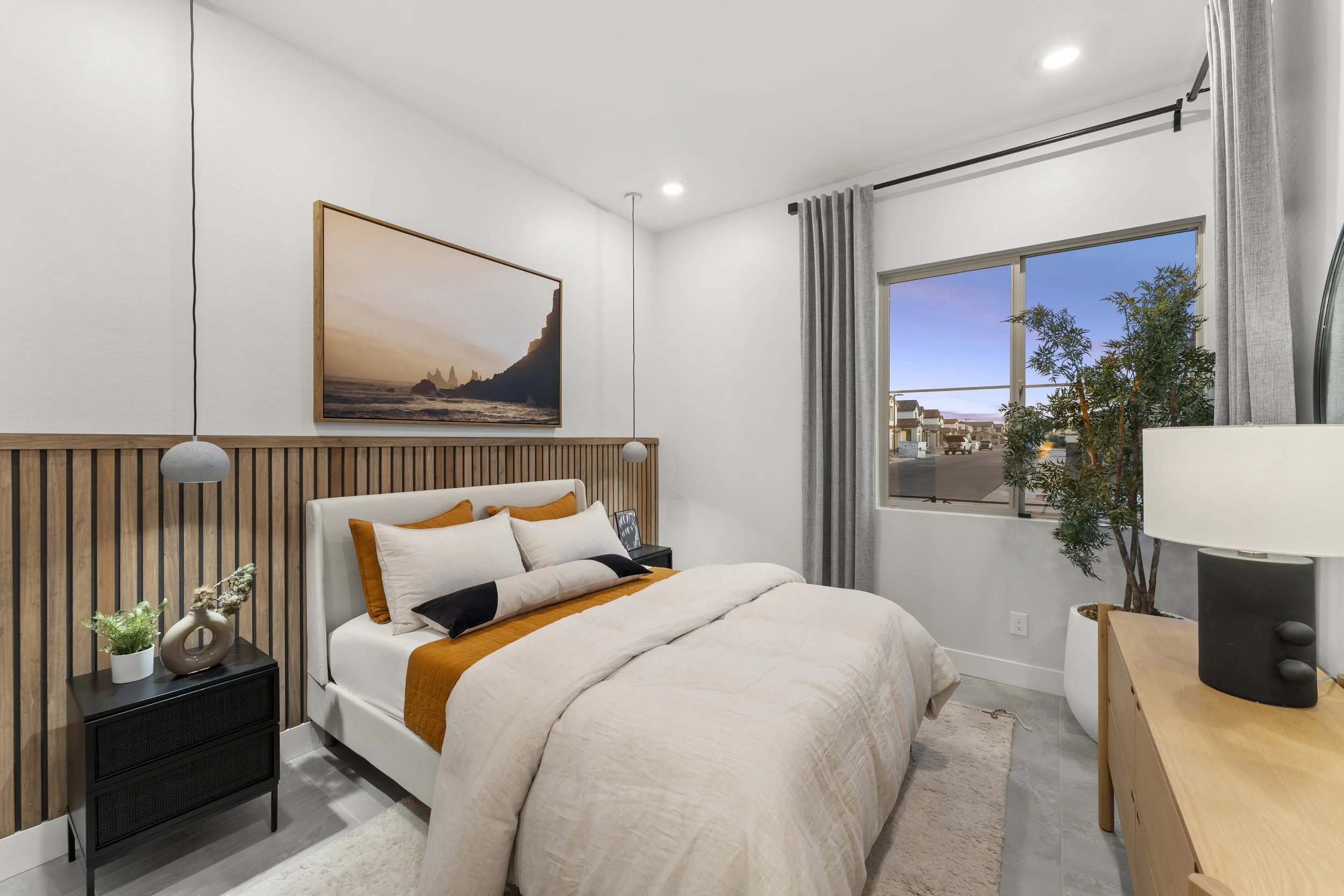 A bedroom with a bed featuring white, black, and ochre pillows and bedding, a wooden slat headboard, a black nightstand with a plant, a hanging pendant light, a large landscape photo on the wall, a window with grey curtains, a potted tree, a wooden d