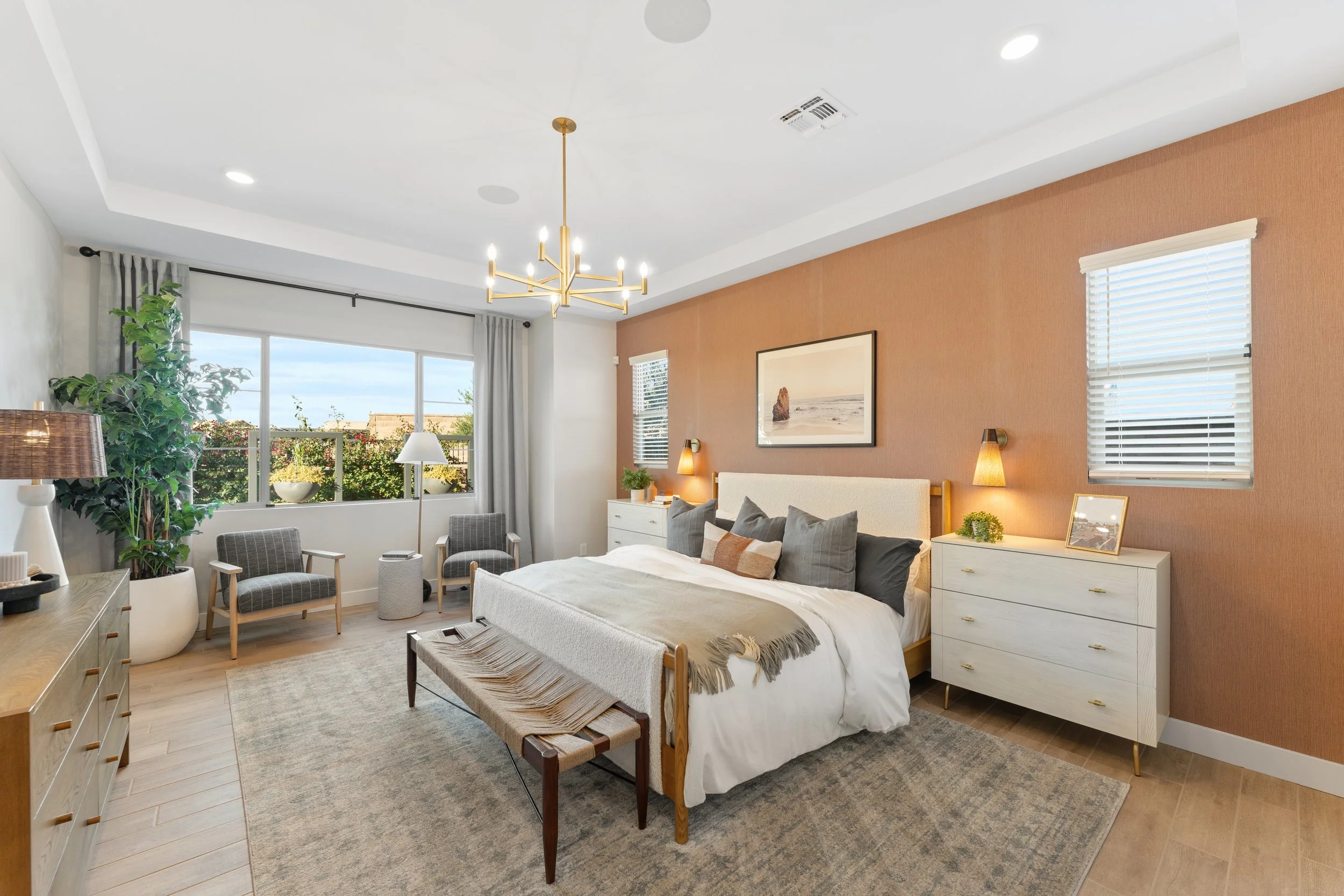 A modern bedroom with a white bed, multiple pillows, and a throw blanket. There are two white dressers with lamps and decor, two windows with blinds, a large sliding window with curtains, a bench at the foot of the bed, an area rug, and seating area 