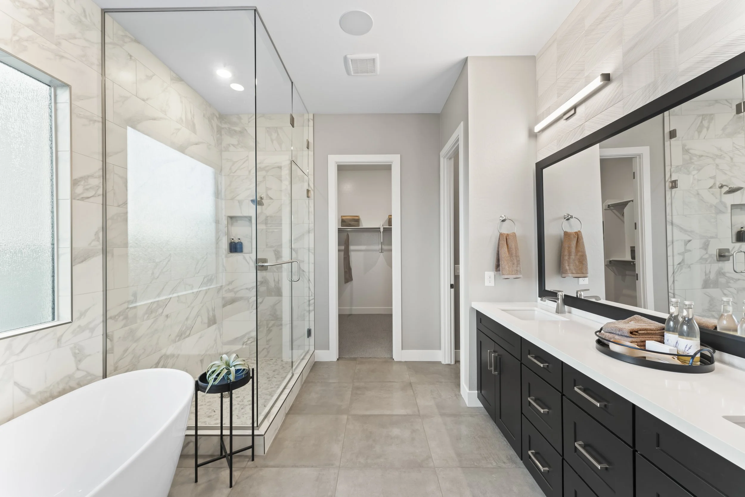 Modern bathroom with a white freestanding bathtub, a glass-enclosed shower with marble tiles, a black vanity with a white countertop, a large mirror, and beige towels.