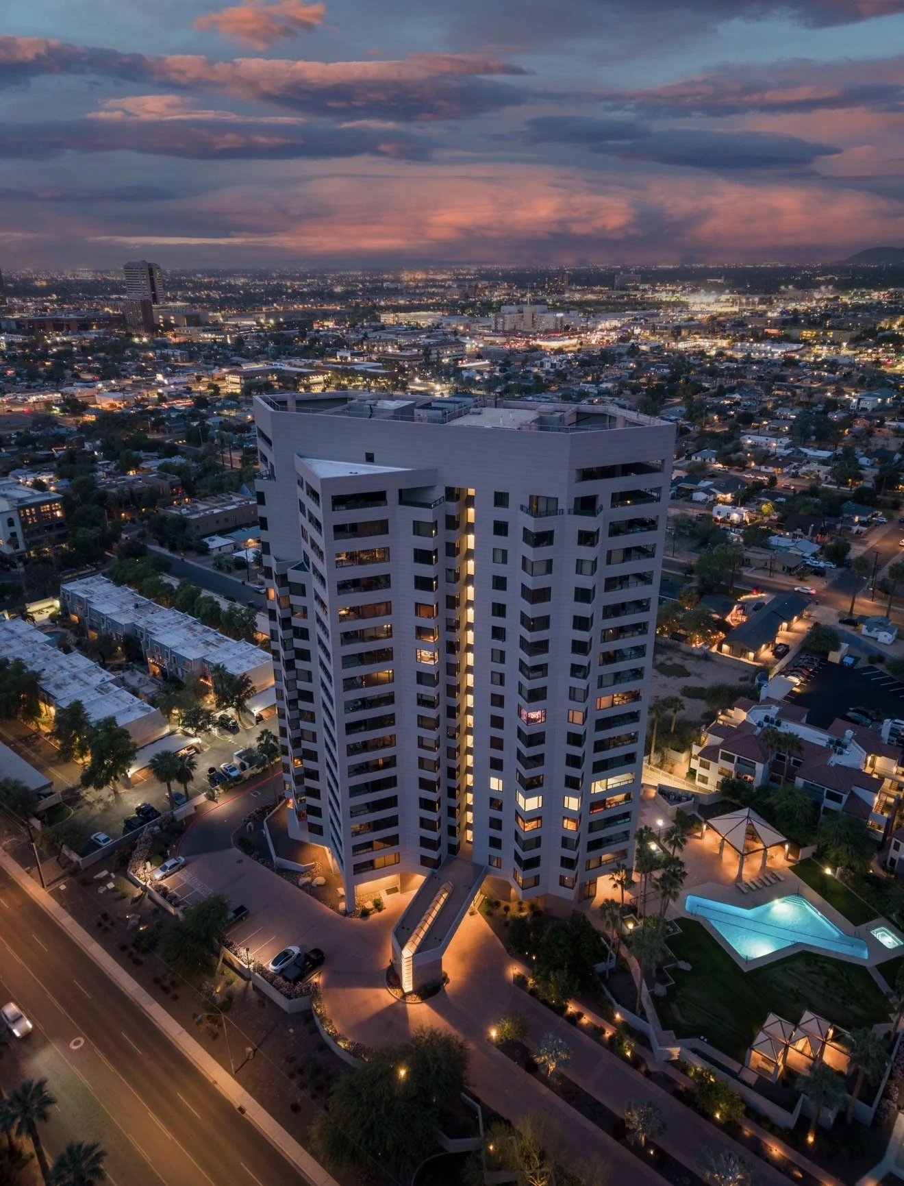 An aerial view of a modern high-rise apartment building at dusk, overlooking a cityscape with a cloudy sky and a pink-orange sunset in the background. The building has outdoor lighting and a swimming pool nearby.
