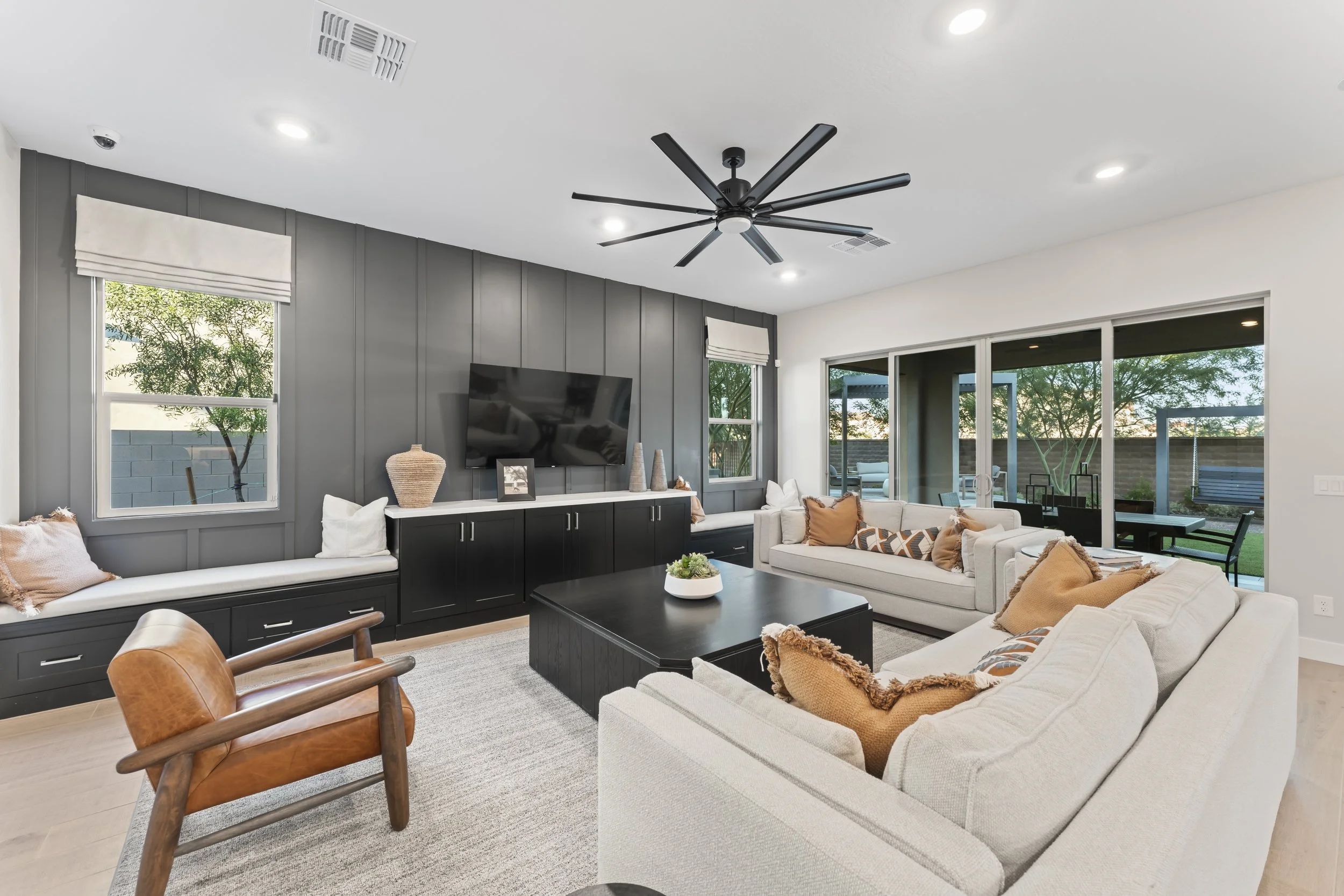 Bright living room with white sofas, a black entertainment console with a flat-screen TV, and a large sliding glass door leading to an outdoor patio with trees.