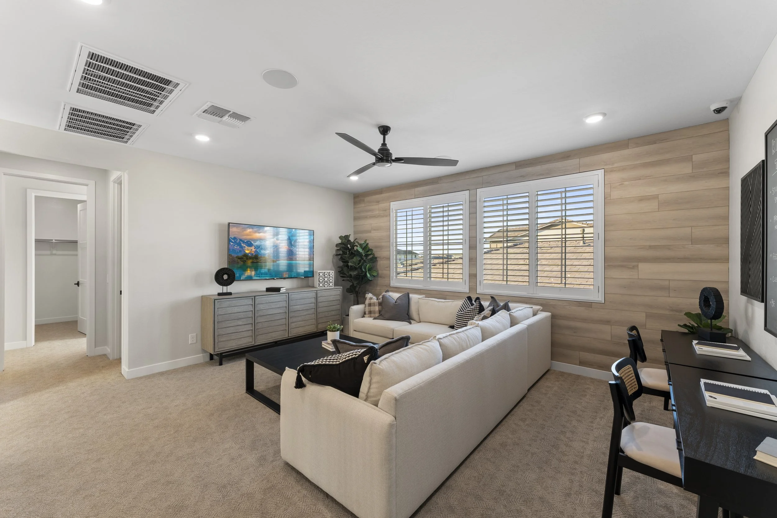 Living room with beige carpet, white walls, and a light wood accent wall. Features a white sectional sofa with black and gray pillows, a black coffee table, a TV on a wooden console, a green plant, and three windows with white blinds. There is a blac