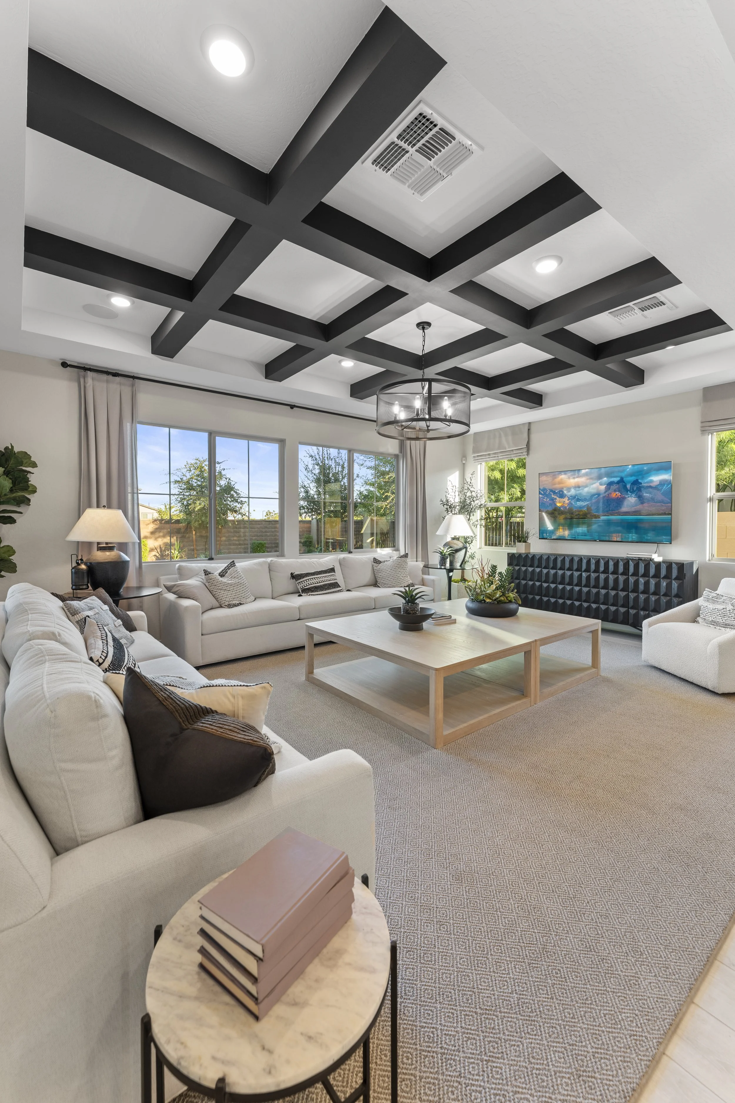 A modern living room with white sofas, a wooden coffee table, a wall-mounted television, large windows, and a coffered ceiling with black beams, decorated with pillows, plants, and lamps.