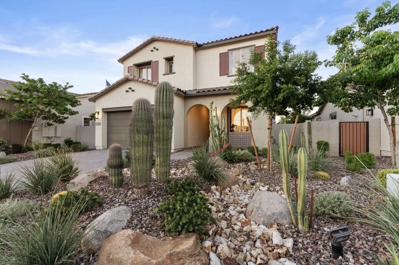 Front yard of a beige two-story house with a garden featuring various cacti and desert plants, a concrete driveway, and a small tree, during twilight.
