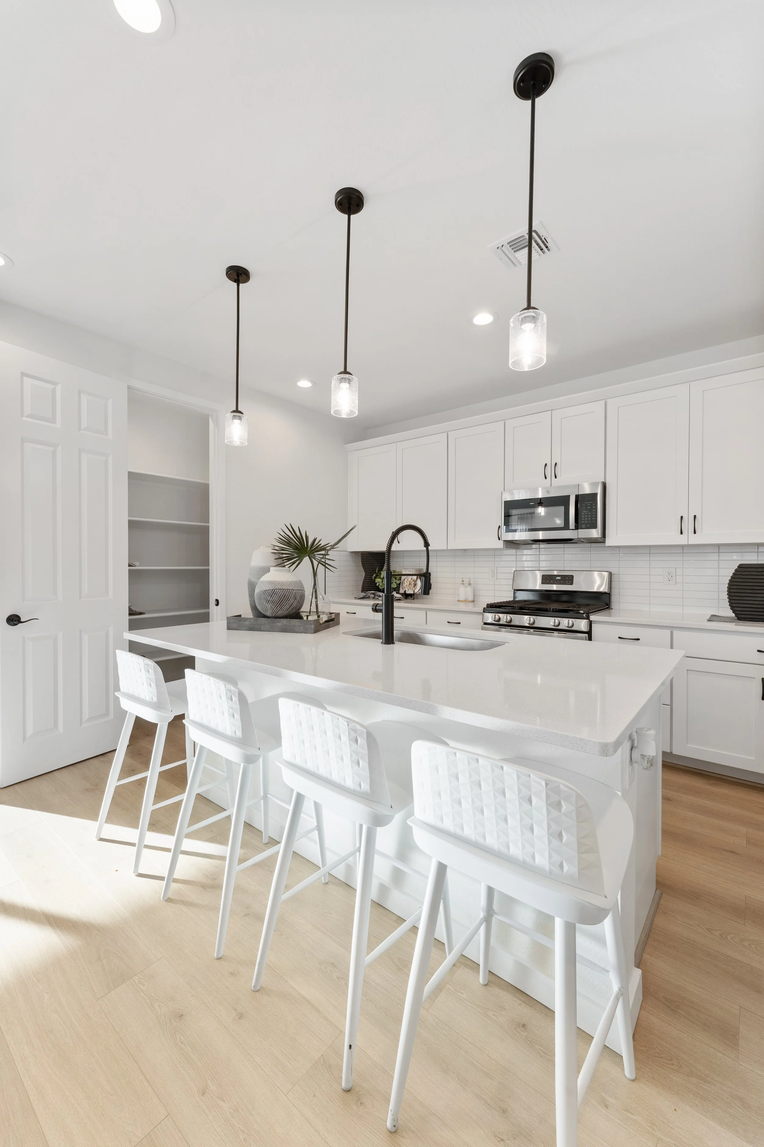 Modern white kitchen with island, four white barstools, pendant lighting, stainless steel appliances, and light wood floors.
